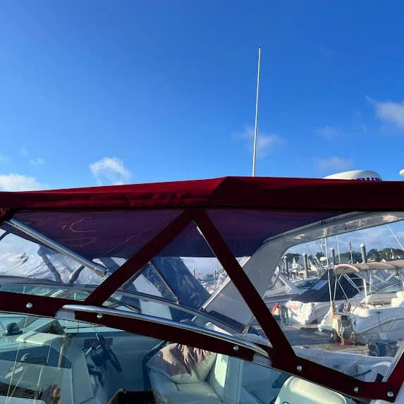 Part of a boat with a red canopy and windows, docked at a marina with other boats in the background, under a blue sky.