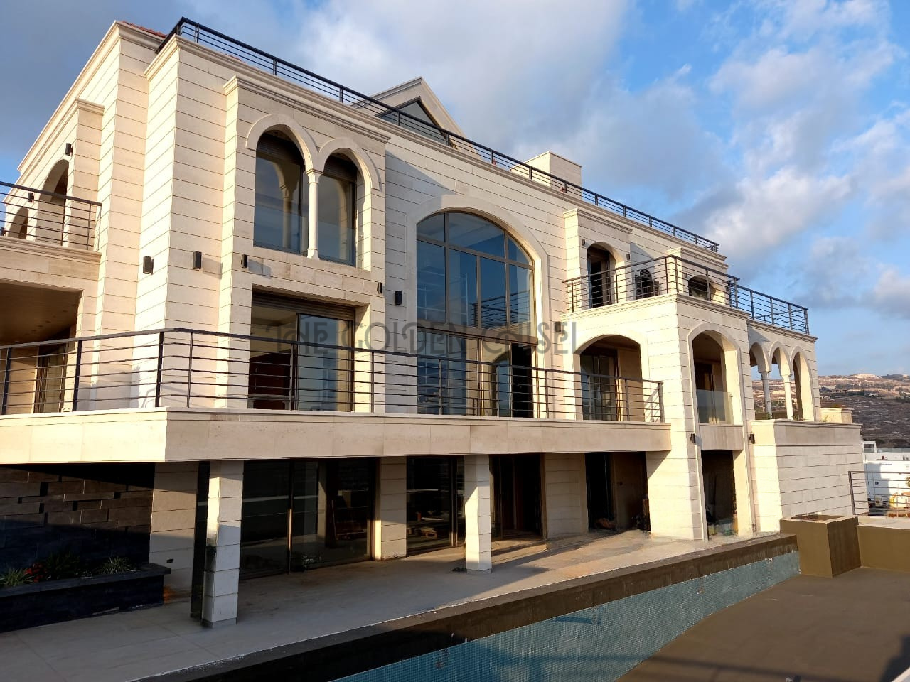 واجهة فيلا فاخرة من الحجر الطبيعي بتصميم عصري وأقواس وشرفات واسعة ونوافذ كبيرة، Luxury modern stone villa exterior featuring natural stone cladding, elegant arches, spacious balconies, and large windows.