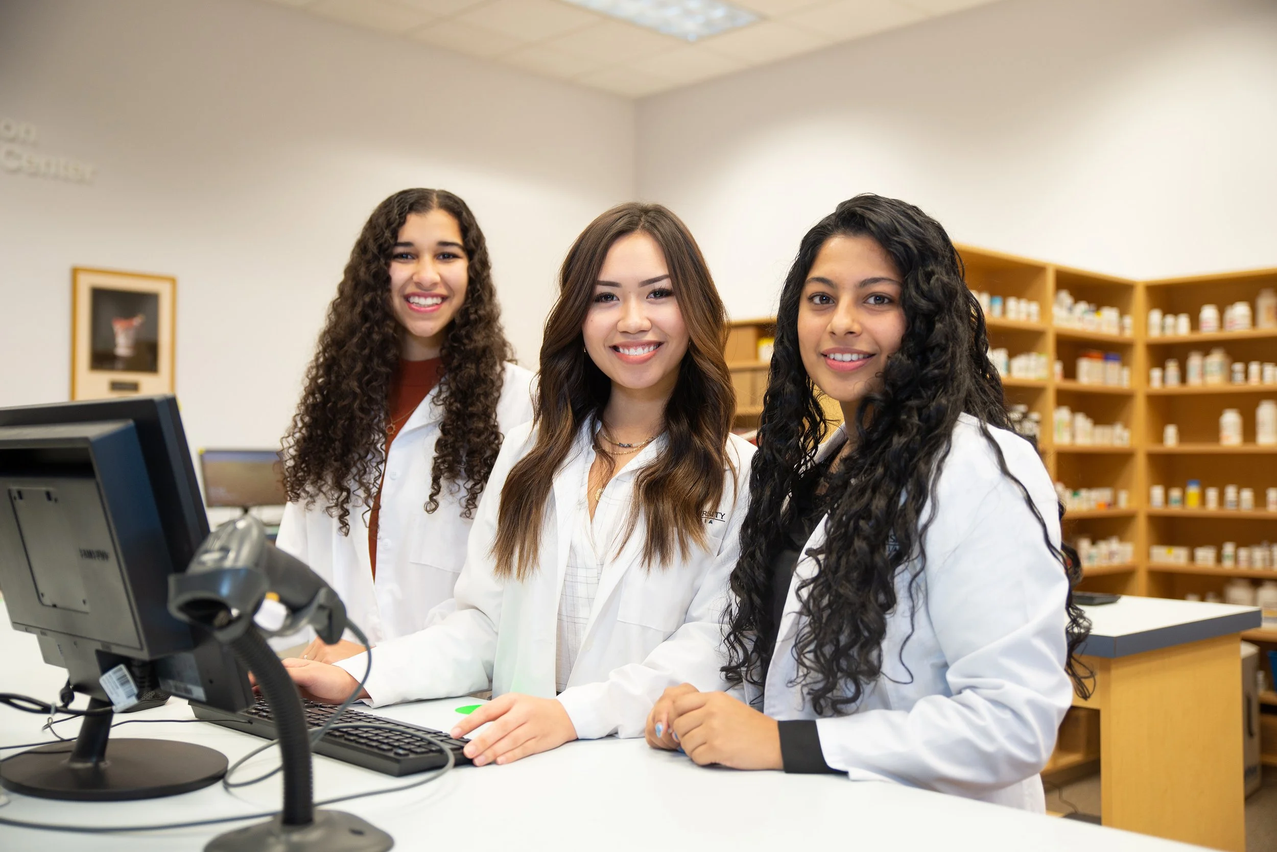 Three young women in white lab coats working at a computer in a laboratory or pharmacy, smiling at the camera.