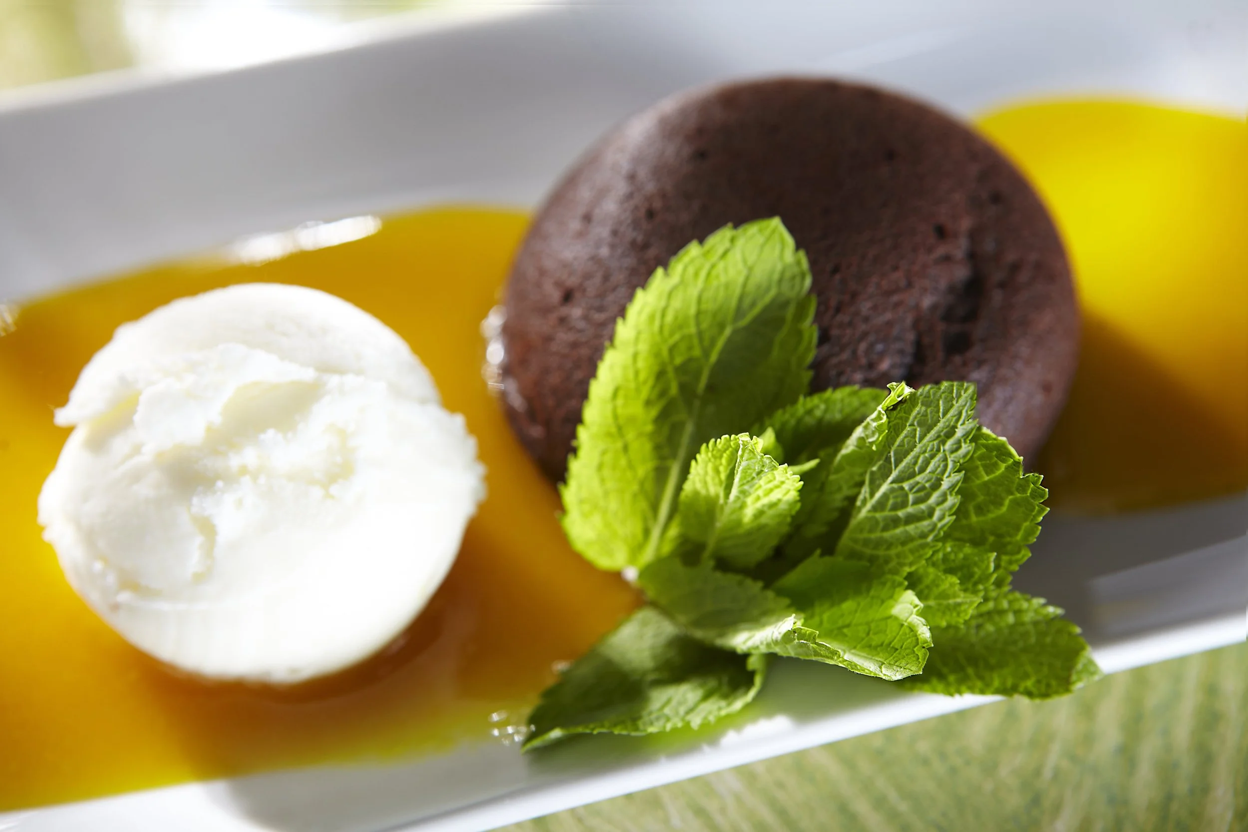 A dessert plate with a scoop of vanilla ice cream, a chocolate cake or cookie, fresh mint leaves, and yellow sauce.