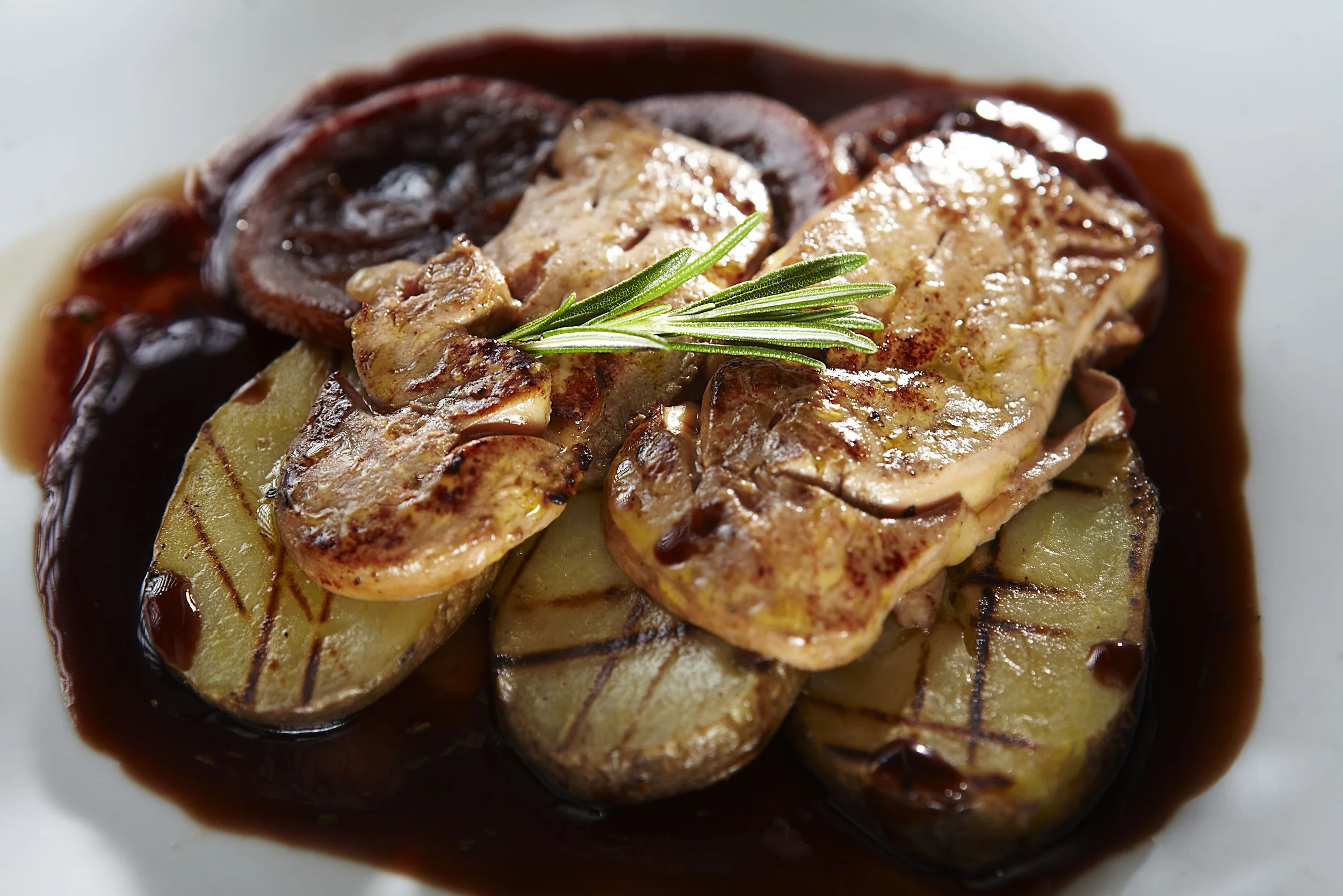 Grilled potato slices topped with grilled mushrooms, sliced beef, and garnished with a sprig of rosemary in a dark sauce.