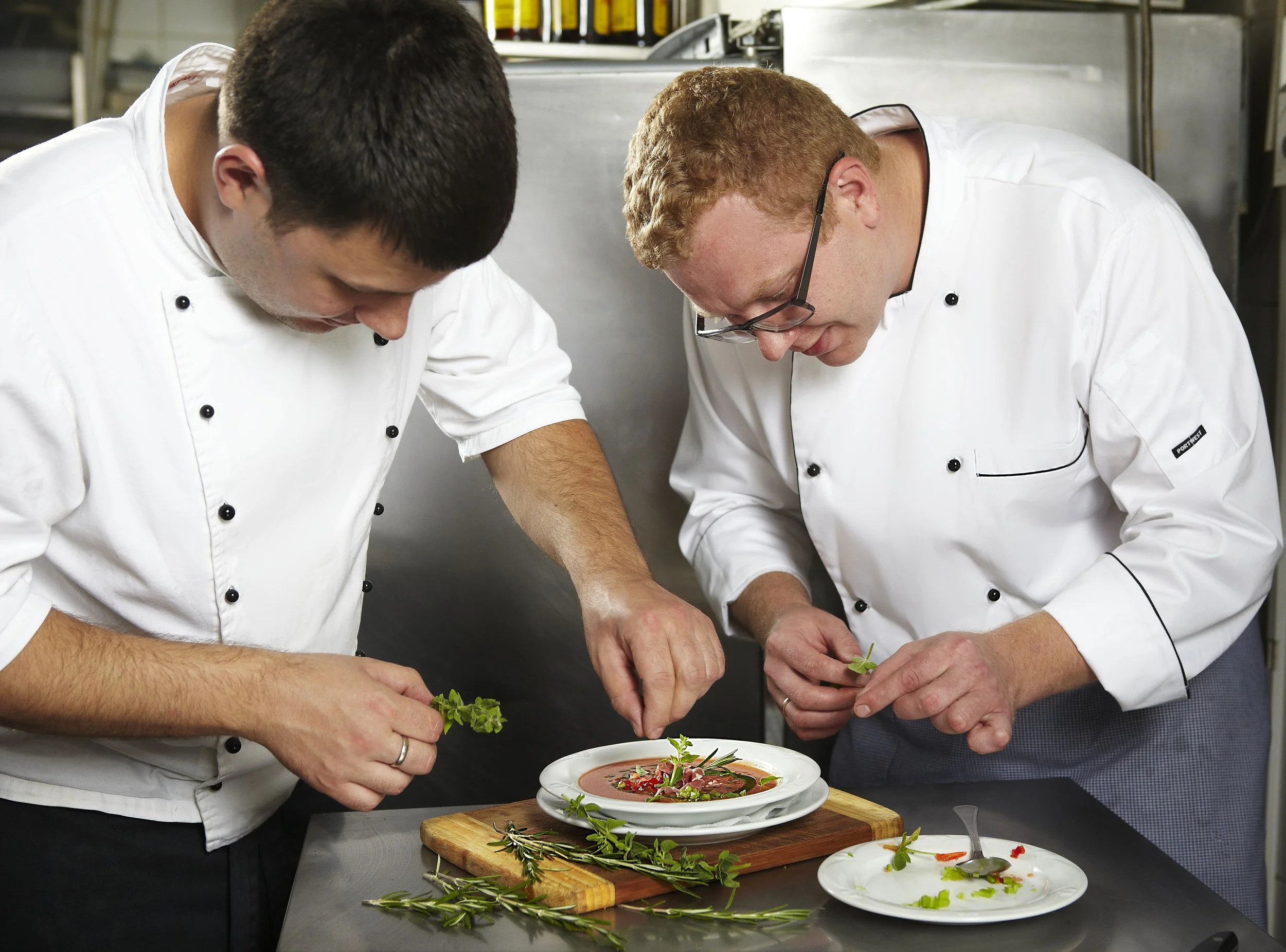 Two chefs in white uniforms garnishing a plated dish with fresh herbs in a professional kitchen.