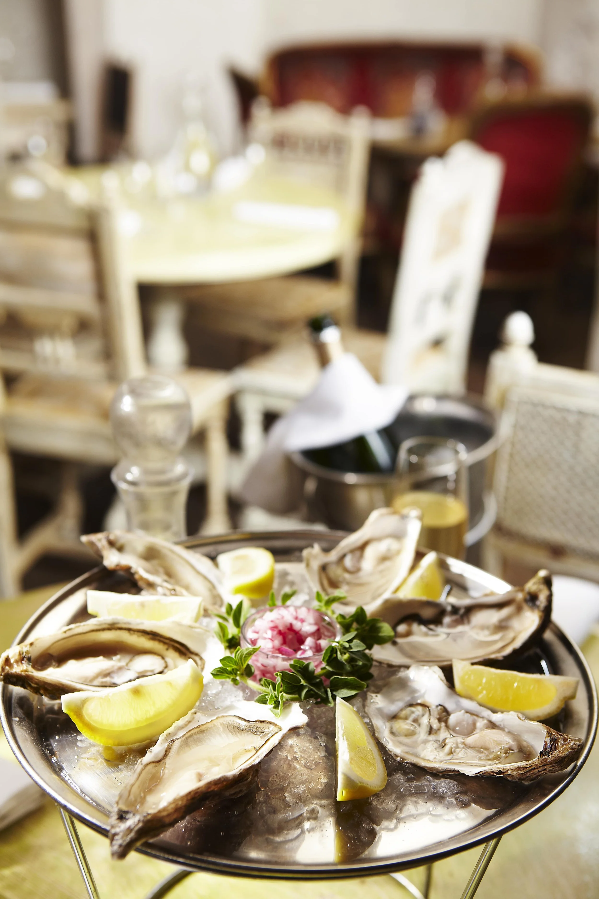 A plate of oysters on ice garnished with lemon wedges and a pink flower in the center, set on a table with a background of a restaurant dining area.