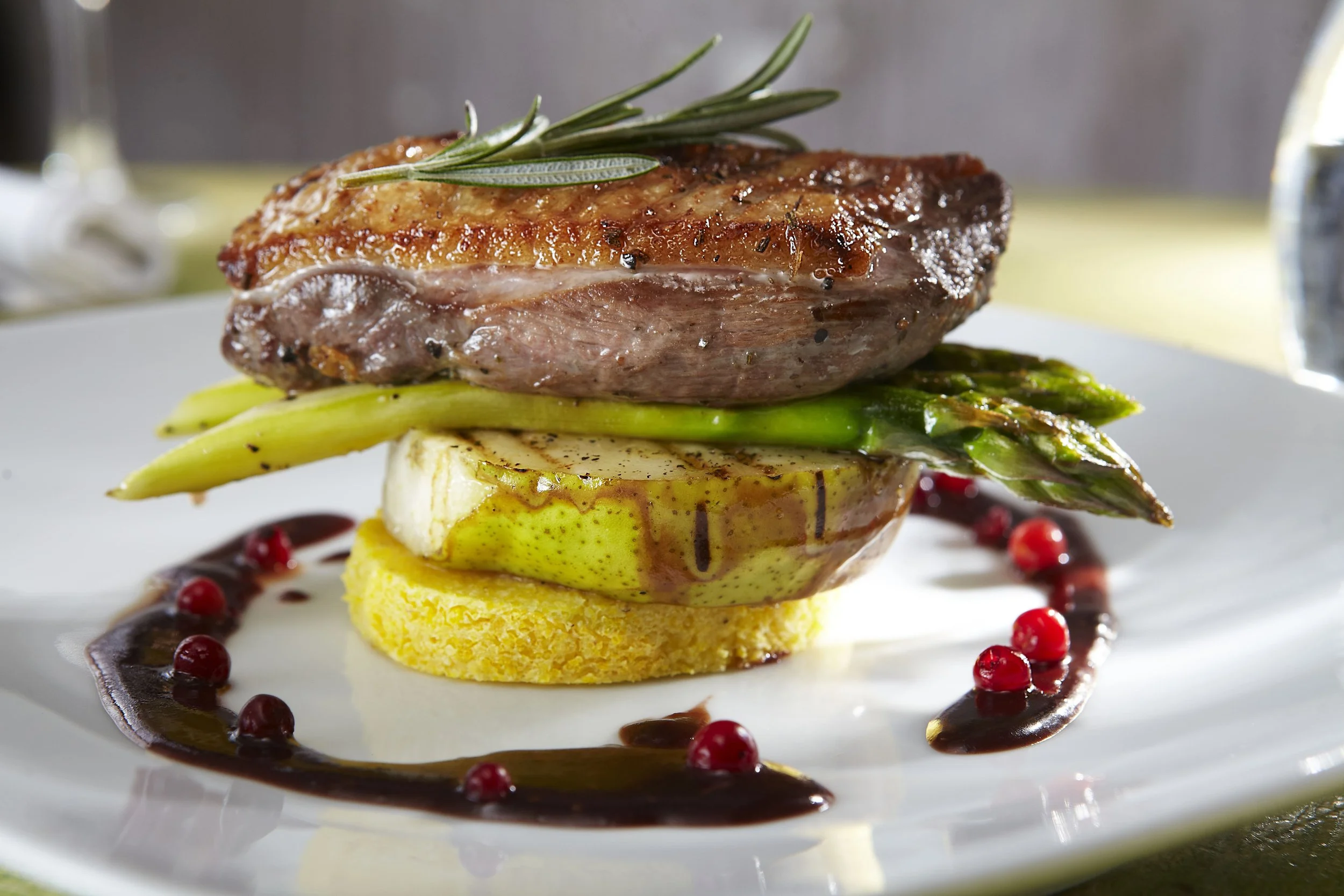 A gourmet dish featuring a cooked steak topped with a sprig of rosemary, served with asparagus, a slice of pear and cheese, on a bed of polenta, garnished with sauce and red berries.