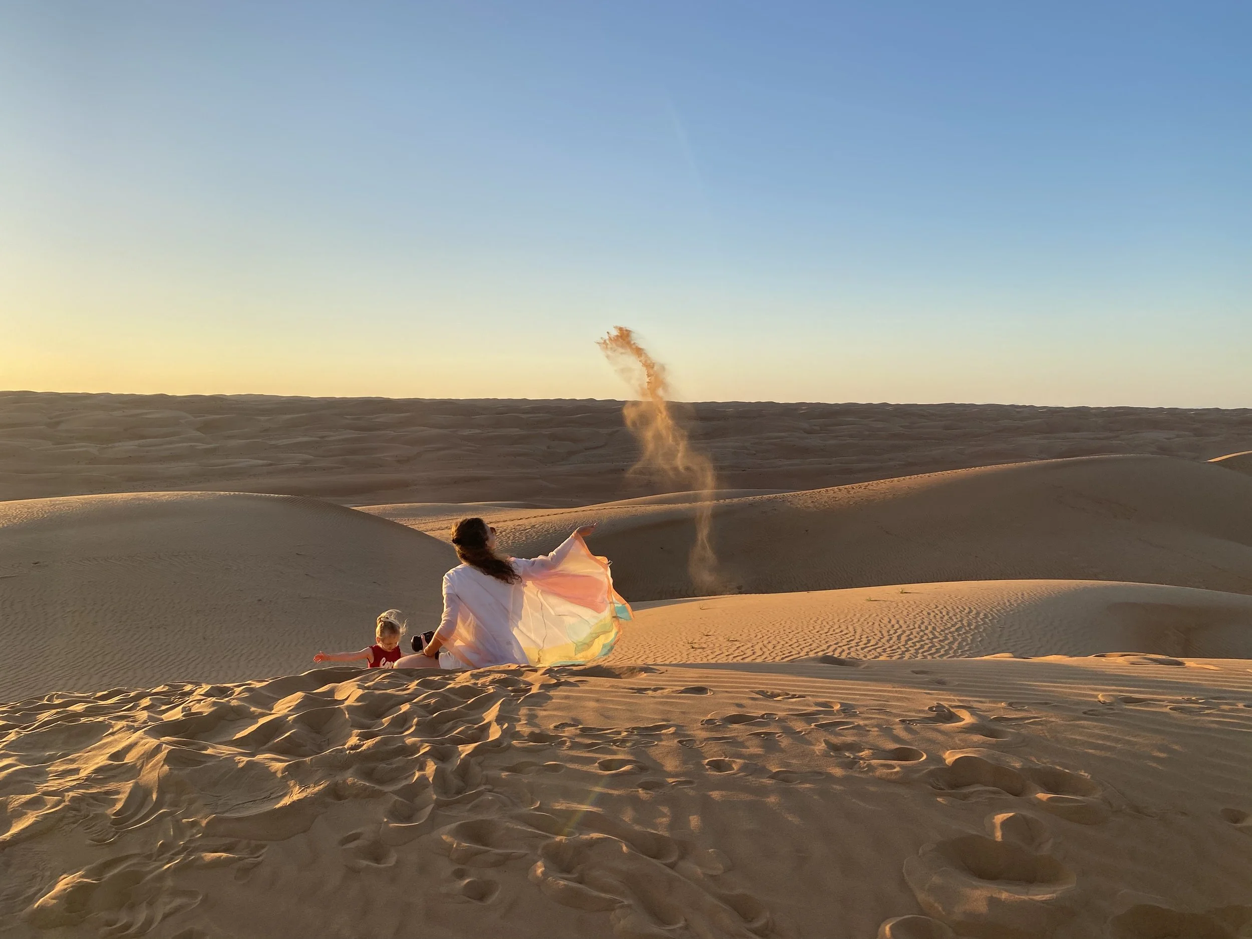 Een vrouw en kind zitten in de woestijn tijdens zonsondergang, de vrouw gooit zand in de lucht, het kindje zit ernaast.