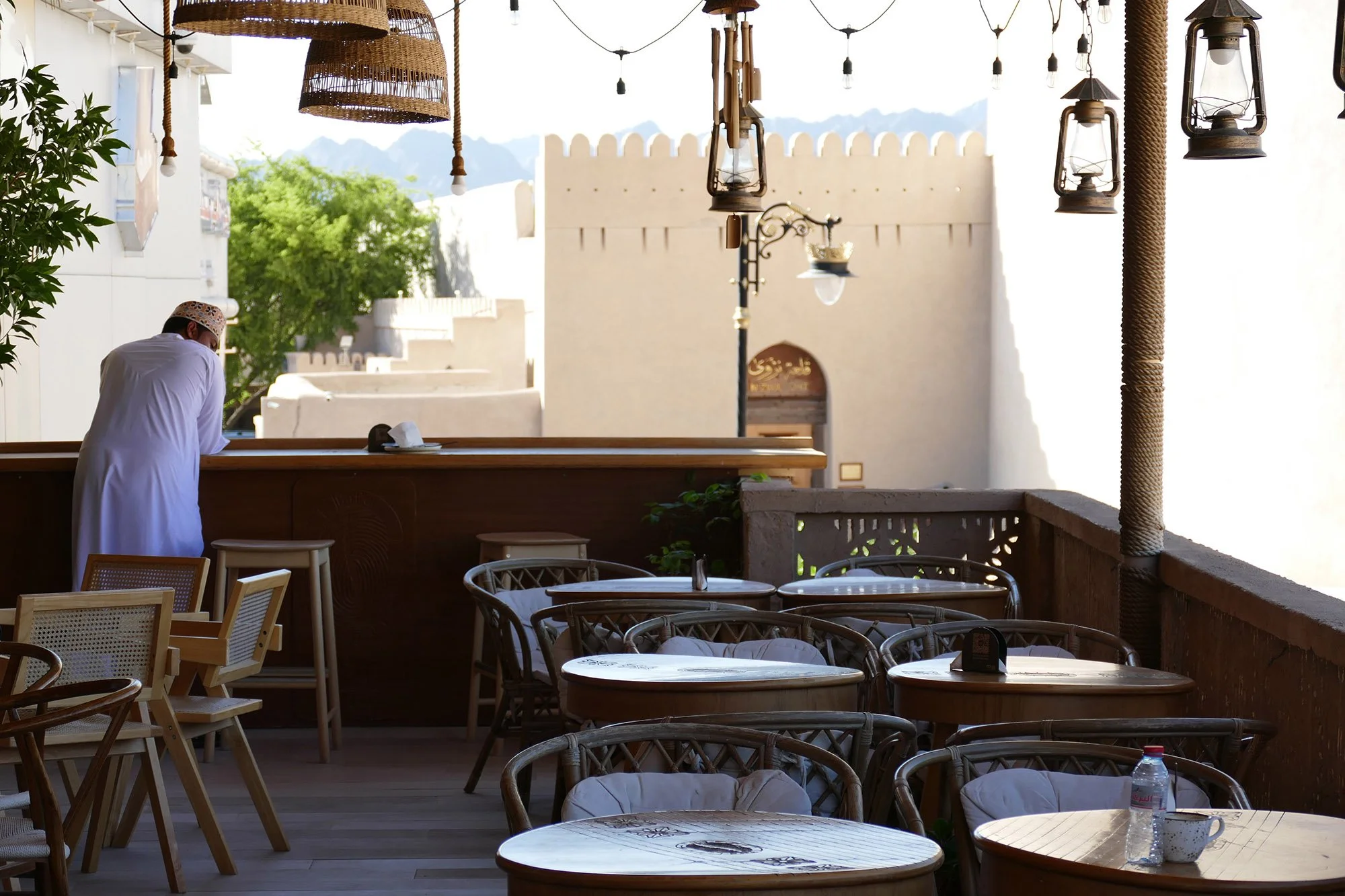 Een dakterras van een restaurant met ronde en vierkante tafels en rieten stoelen. In de achtergrond is een man te zien die een wit gewaad en een hoofddeksel draagt, met uitzicht op een ommuurde stad met bergen op de achtergrond.