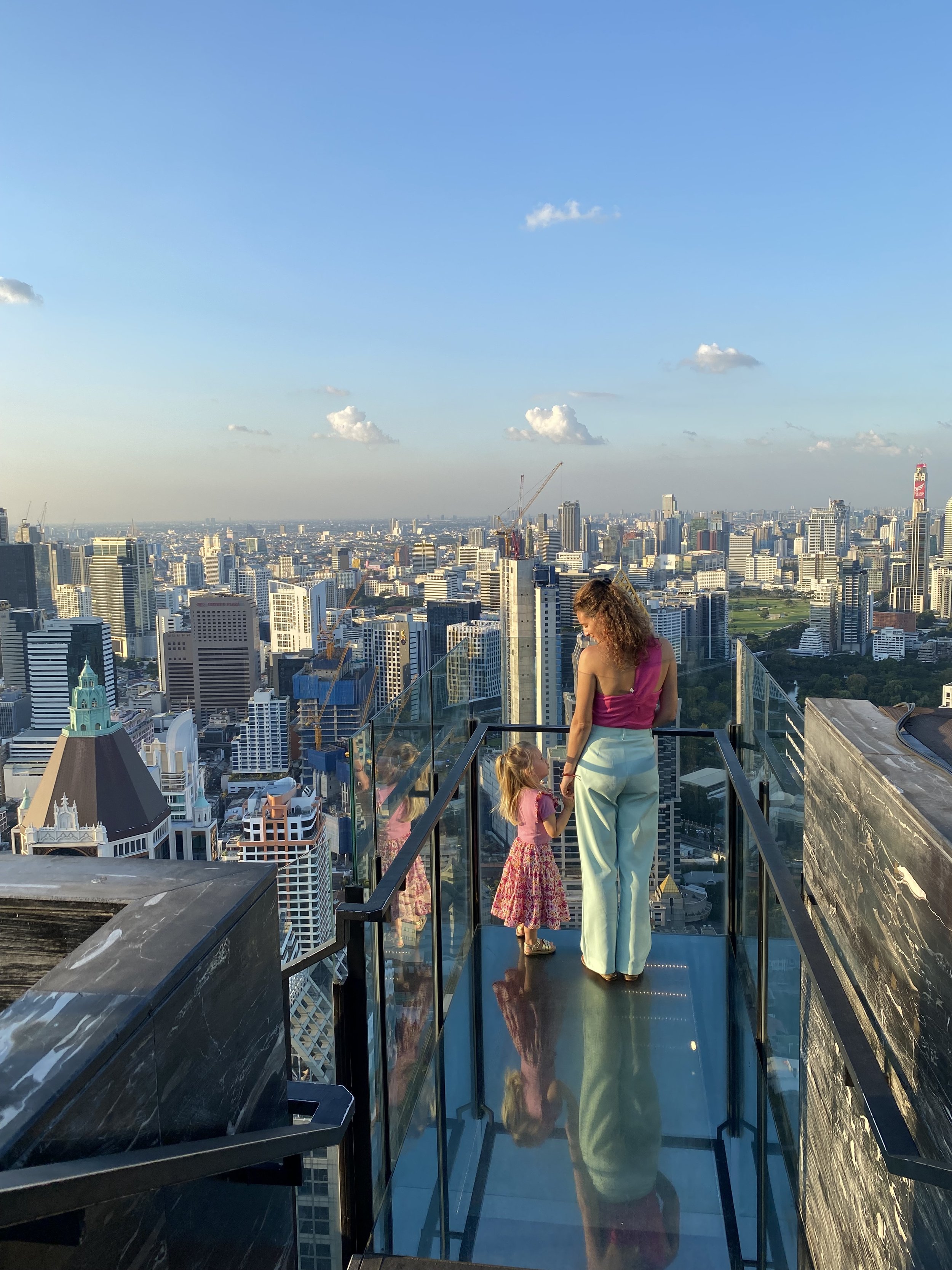 Een vrouw en twee jonge meisjes op een balkon met uitzicht over een stedelijk skyline met hoge gebouwen en wolken, onder een blauwe hemel.