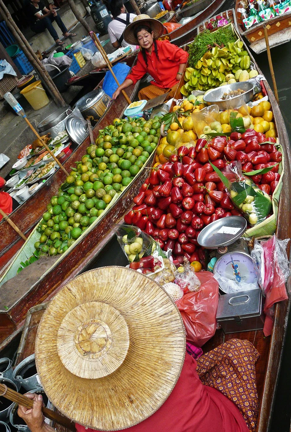 Vrouw verkoopt verse fruit op een marktkraam met ramekins en weegschalen, omgeven door rode, groene en gele fruitsoorten.
