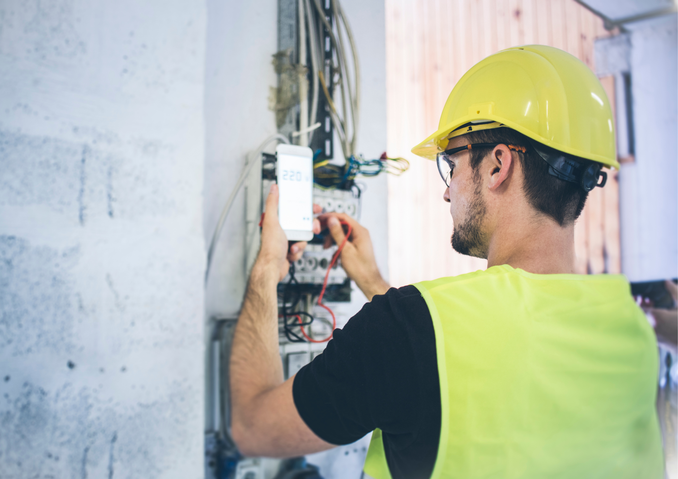A construction worker wearing a yellow safety helmet and vest working on electrical wiring using a multimeter or testing device.