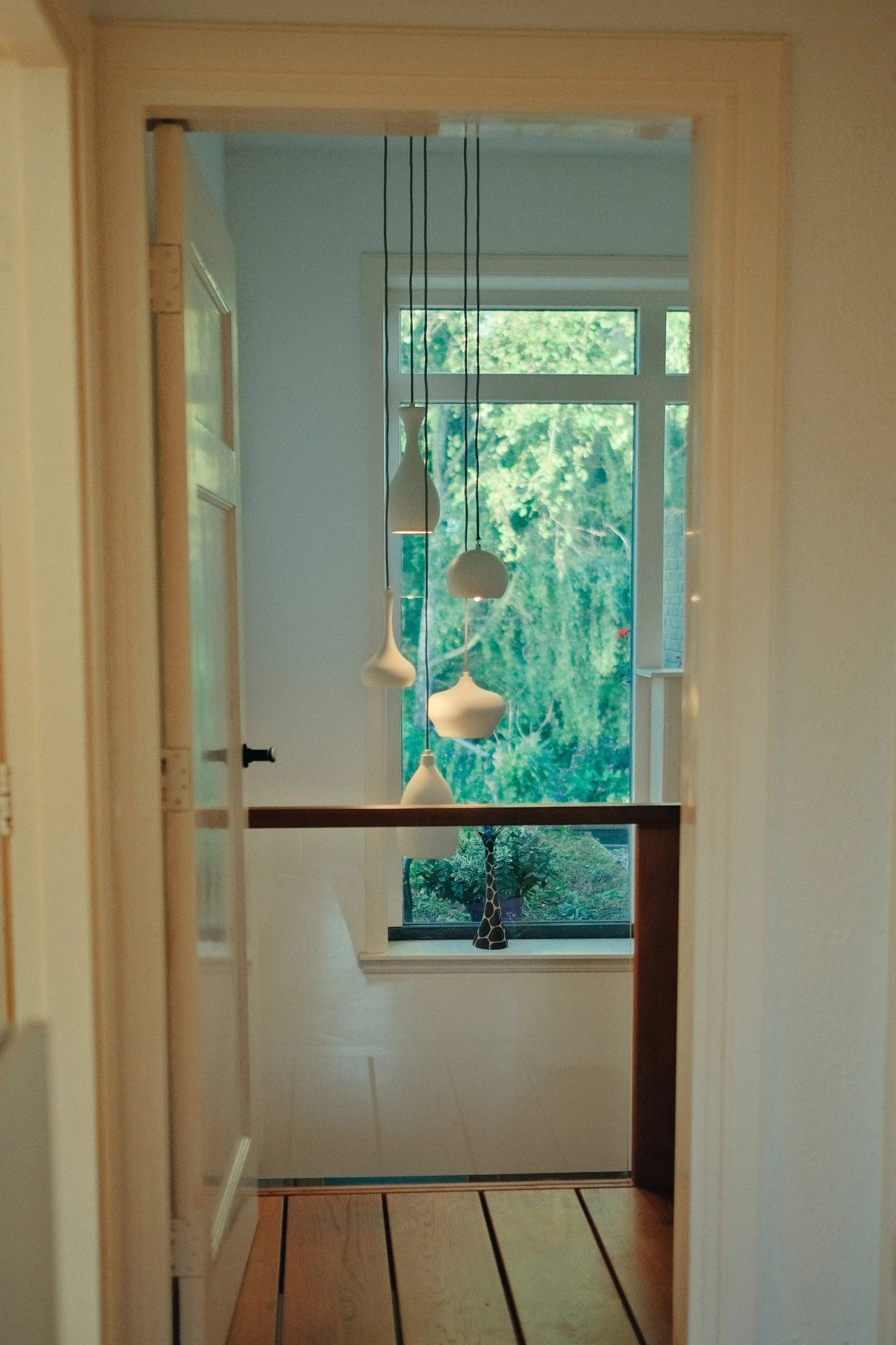 Interieur van een kamer gezien door een deuropening, met witte hanglampen, een raam met groen uitzicht, en een opvallend decoratief giraffenbeeld op de vensterbank.