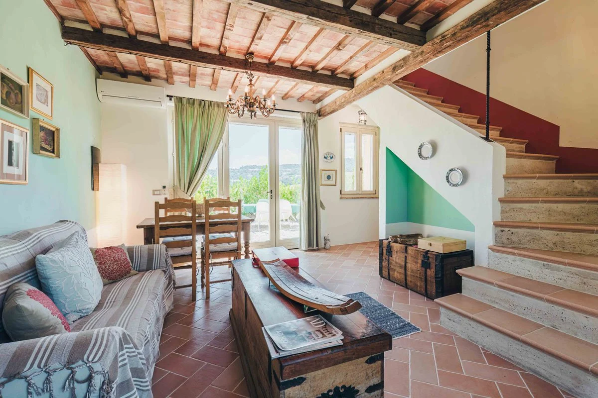Living area of the Casa Janna Cottage with exposed wooden beams, dining table, sofa, staircase, and glass doors opening outside