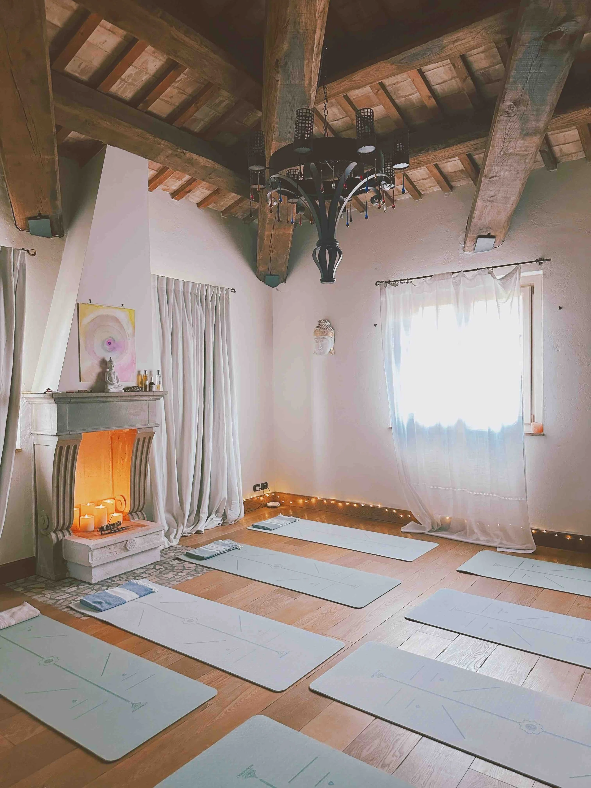 Yoga studio with white mats laid out on wooden floor, white curtains on windows, ceiling with exposed wooden beams, a chandelier, and a fireplace with candles and decorative items.