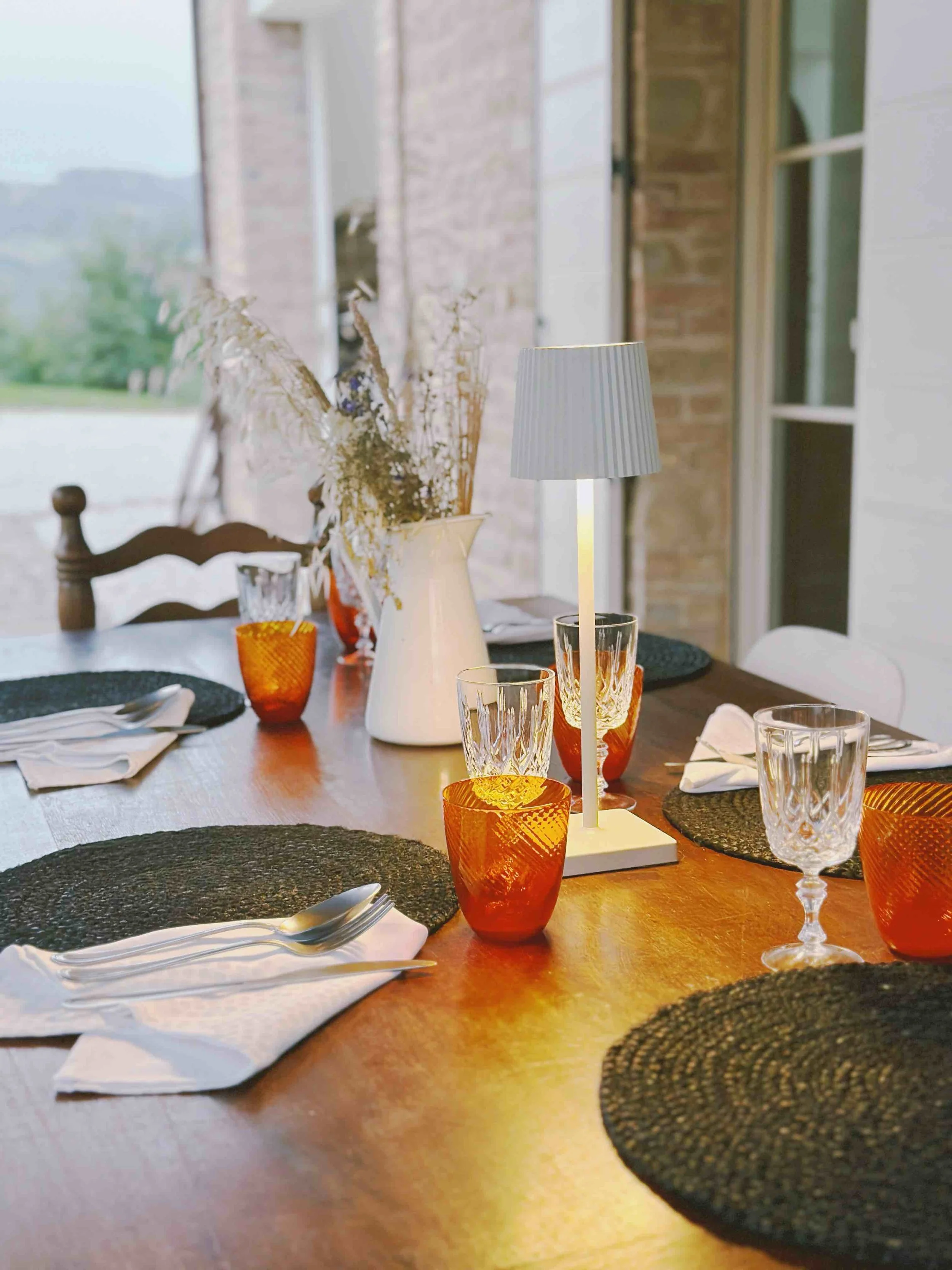A cozy dining table set with black woven placemats, white napkins, and glassware featuring goblets and orange textured cups. A white pitcher with dried flowers and a modern table lamp add to the decor, with a large window in the background letting in natural light.