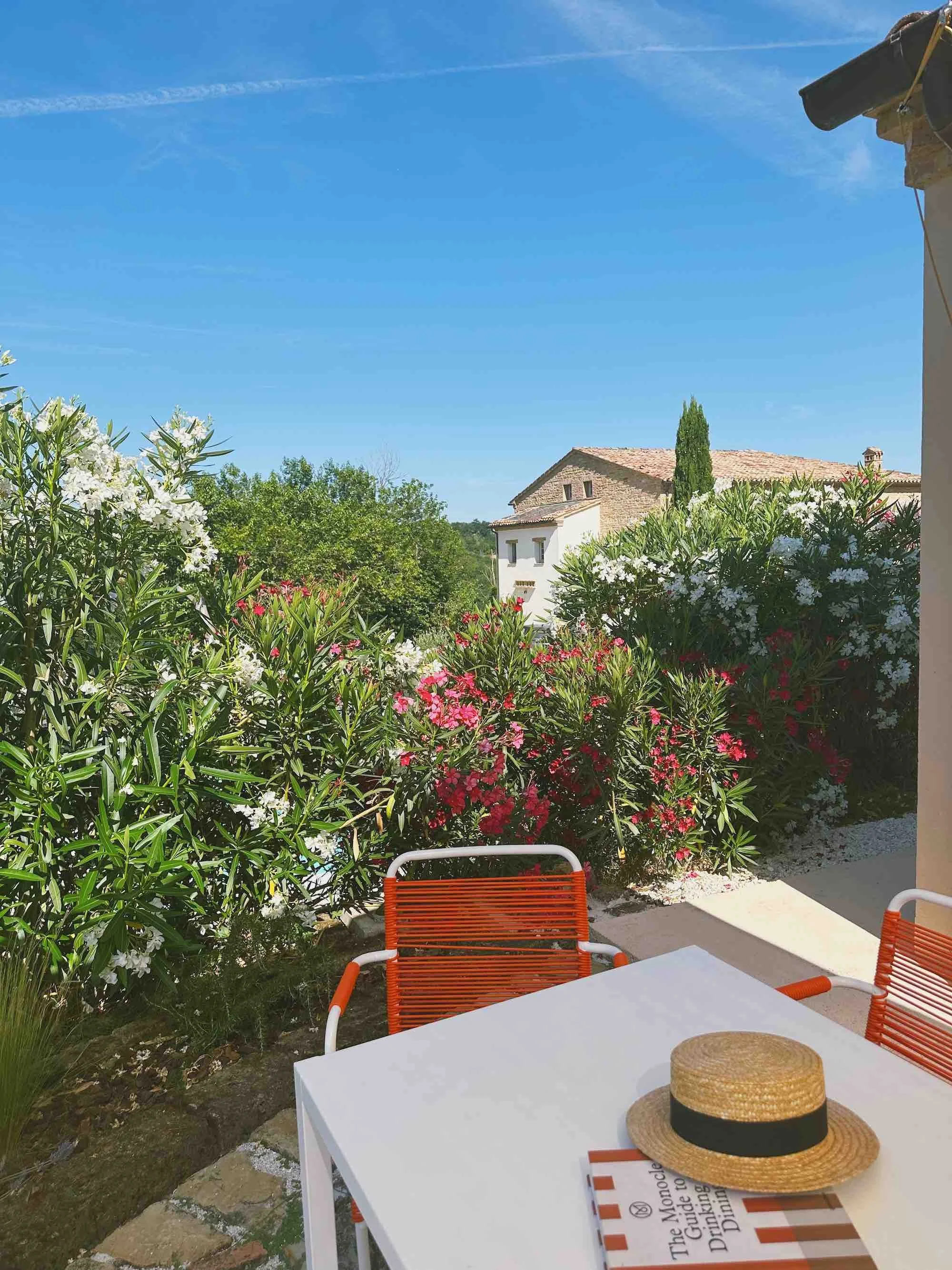 Outdoor patio table with a straw hat and a book, surrounded by lush flowering bushes and a clear blue sky, part of the outdoor Sole Junior Suite of Casa Janna