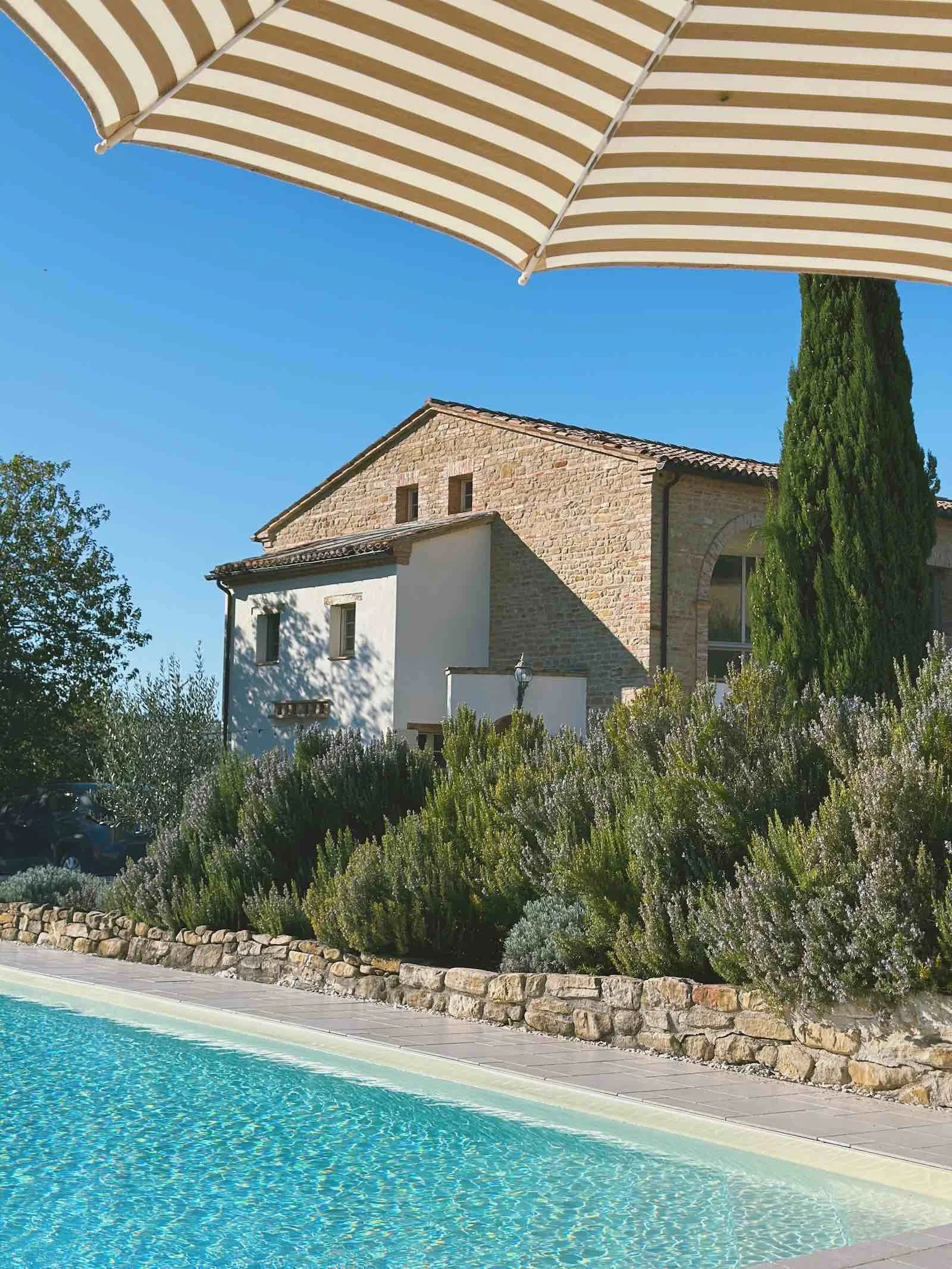 A swimming pool under a beige and white striped umbrella, with a stone house and lush green shrubs and trees in the background.