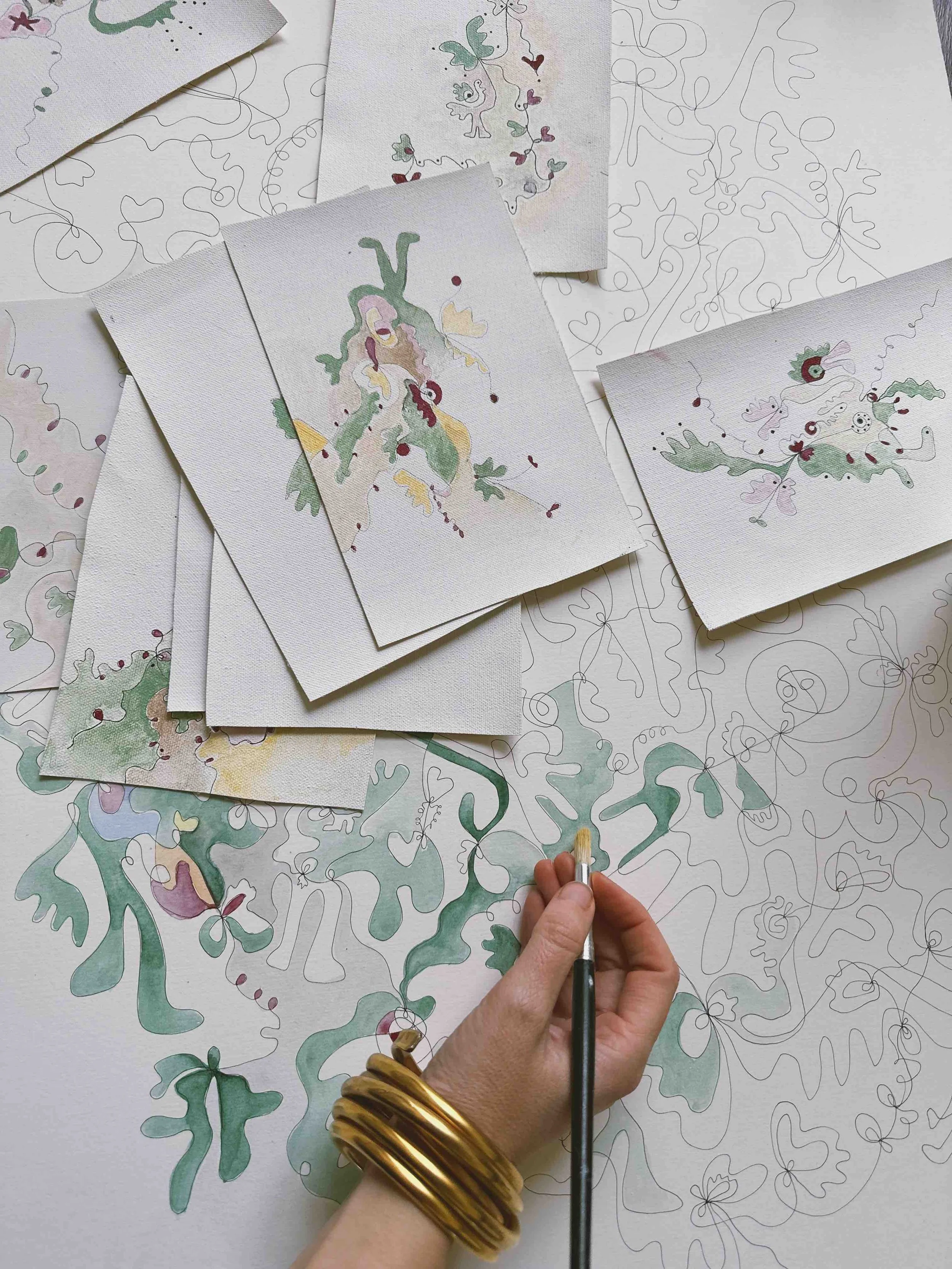Francesca is drawing and painting abstract and whimsical floral and organic shapes on a large sheet of paper, with several smaller finished pieces and sketches of similar designs scattered around.