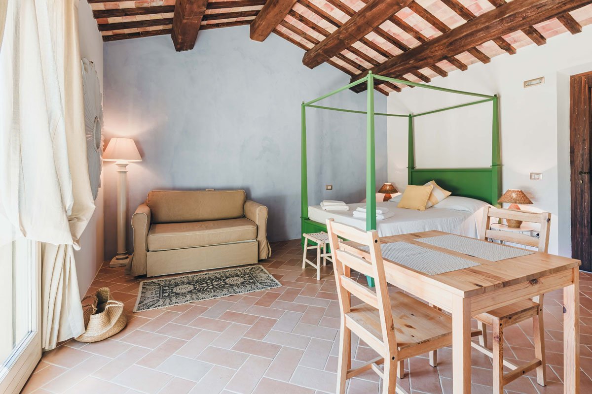 Interior of a sunlit room with a terra-cotta tiled floor, white walls, and exposed wooden beams on the ceiling; includes a beige sofa, a standing lamp, a small area rug, a wooden dining table with four chairs, a bed with a green frame, pillows, and bedside lamps.