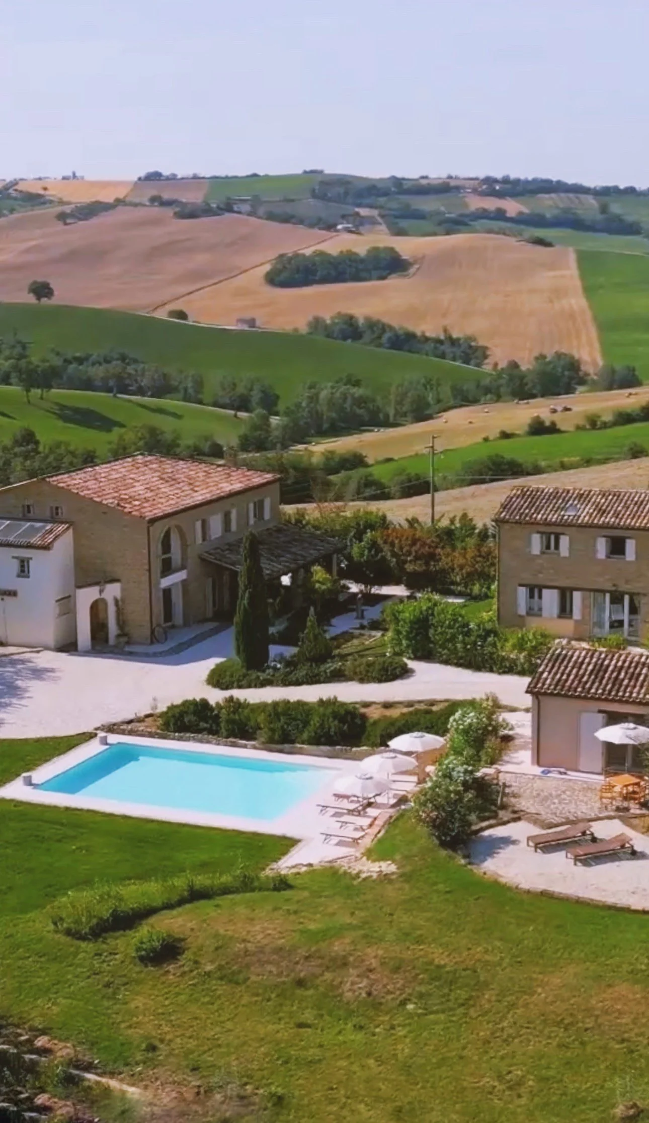 Aerial view of a Mediterranean-style villa - Casa Janna - and swimming pool surrounded by lush greenery and Le Marche rolling hills in the countryside.