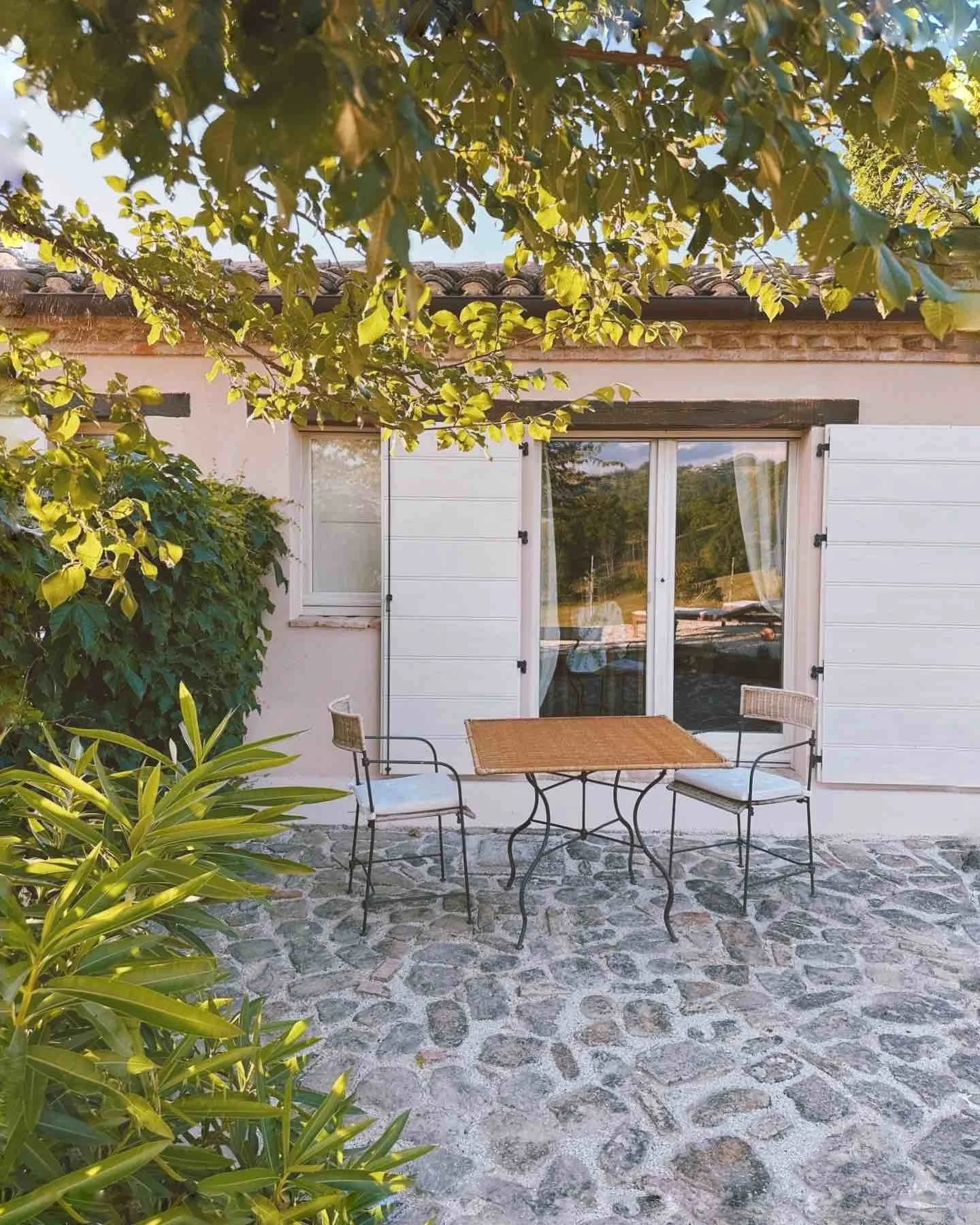 A small outdoor patio with a stone floor, featuring a metal table with a wooden top and two wicker chairs with cushions, in front of a house with white shutters and glass doors, surrounded by greenery and a tree.
