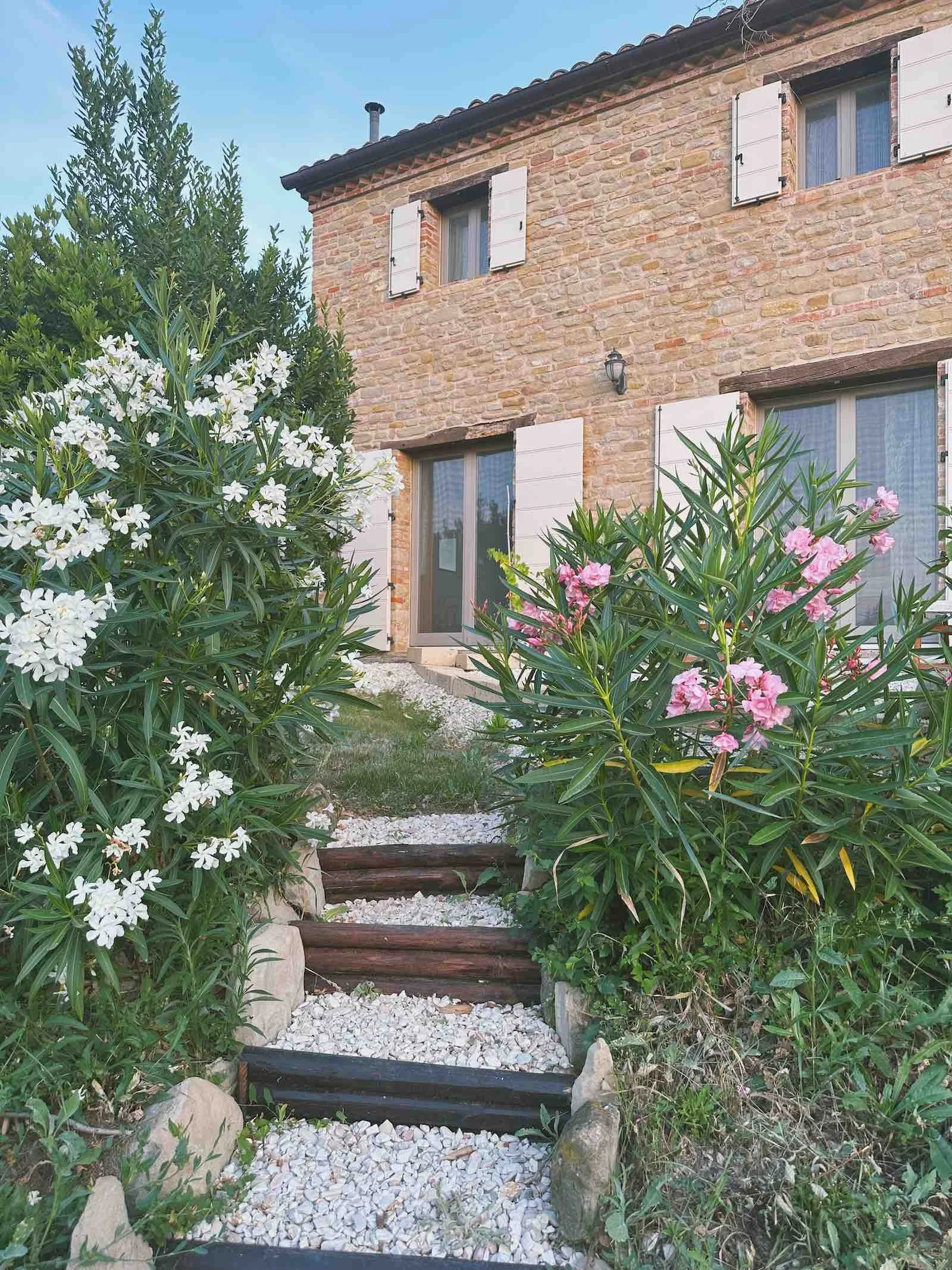 A Stone guesthouse with white shutters at Casa Janna, set in the hills of San Ginesio, Le Marche. Garden path leading to the Casa Janna guesthouse, bordered by flowers and greenery