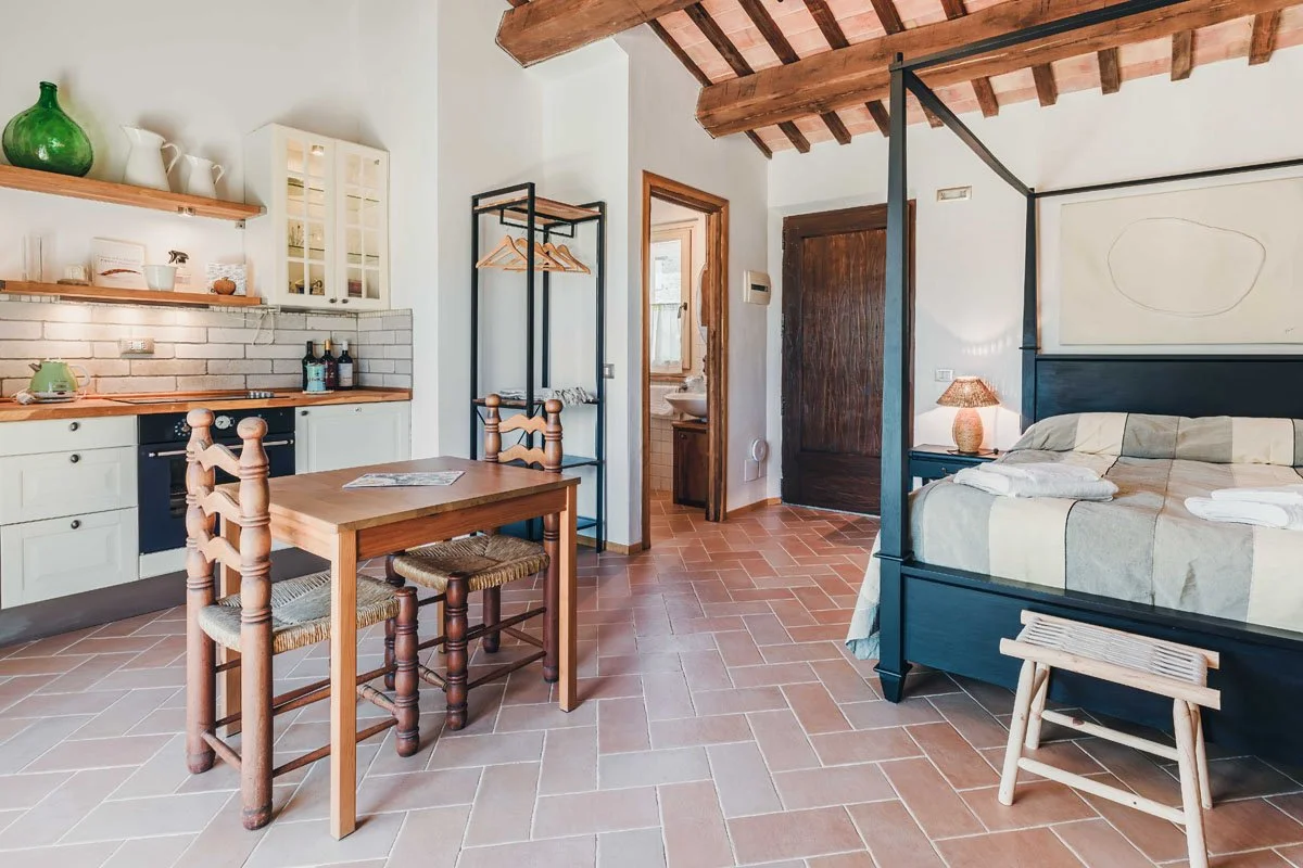 Interior of a cozy studio apartment with a kitchen area, a dining table with three chairs, a bed with linens, and rustic ceiling beams.