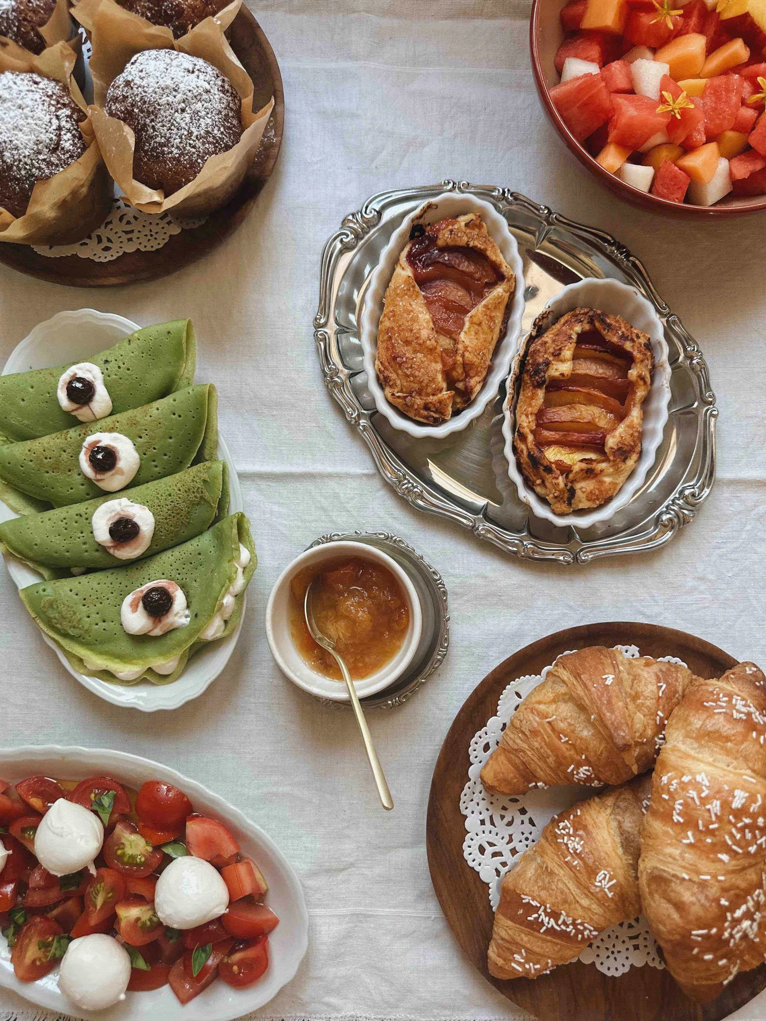 A breakfast table with muffins, croissants, fruit salad, watermelon, and green crepes topped with cream and blackberries.