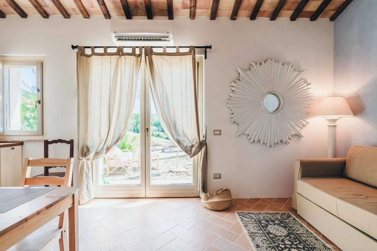 Bright living room with white walls, wooden ceiling, beige curtains on glass doors leading outside, a beige sofa, a large decorative mirror, and a floor lamp with a beige shade.