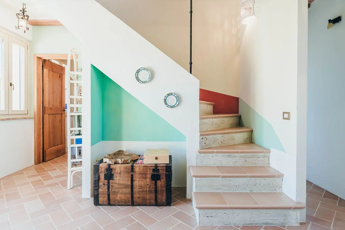 Interiors of The Cottage, Casa Janna entryway with tiled floor, staircase with concrete steps and pink tiles, a wooden door, a window, and decorative plates on a painted wall.