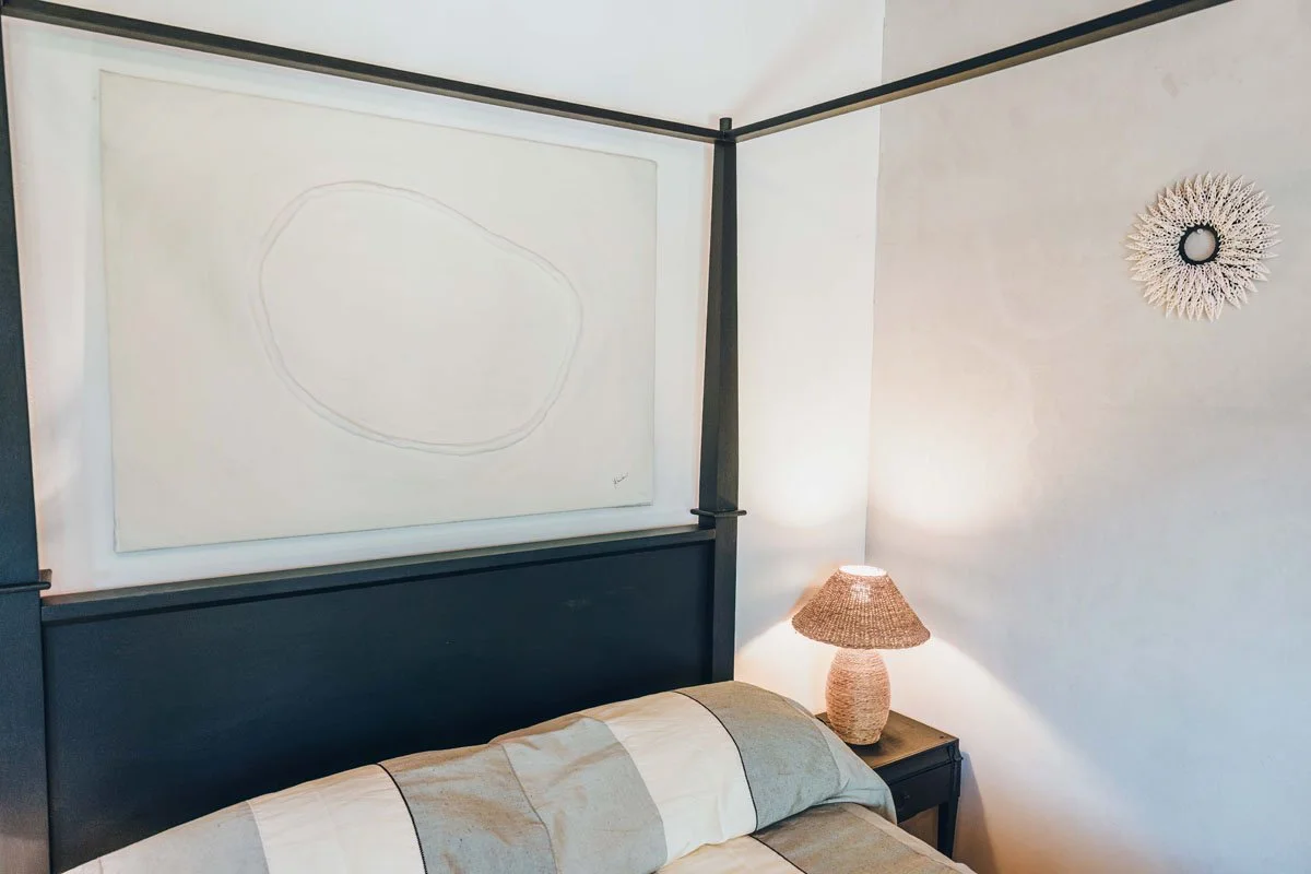 A bedroom corner with a bed, a hanging painting, a bedside table with a lamp, and decorative wall art.