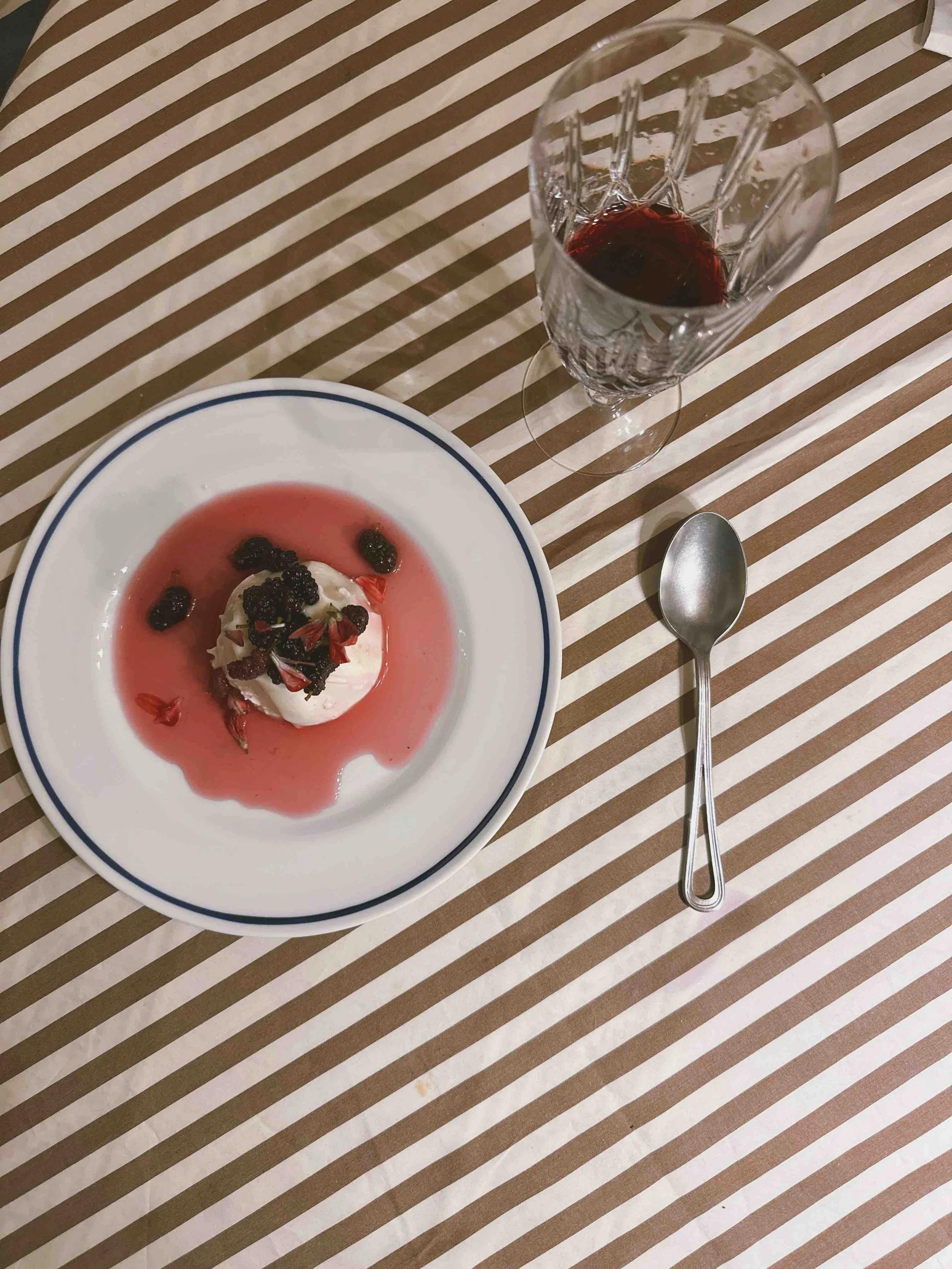 A panacotta topped with berries and edible flowers, filled with pink syrup, on a white plate with a blue rim. A tall glass of vino cotto and a silver spoon are on a brown and white striped tablecloth.