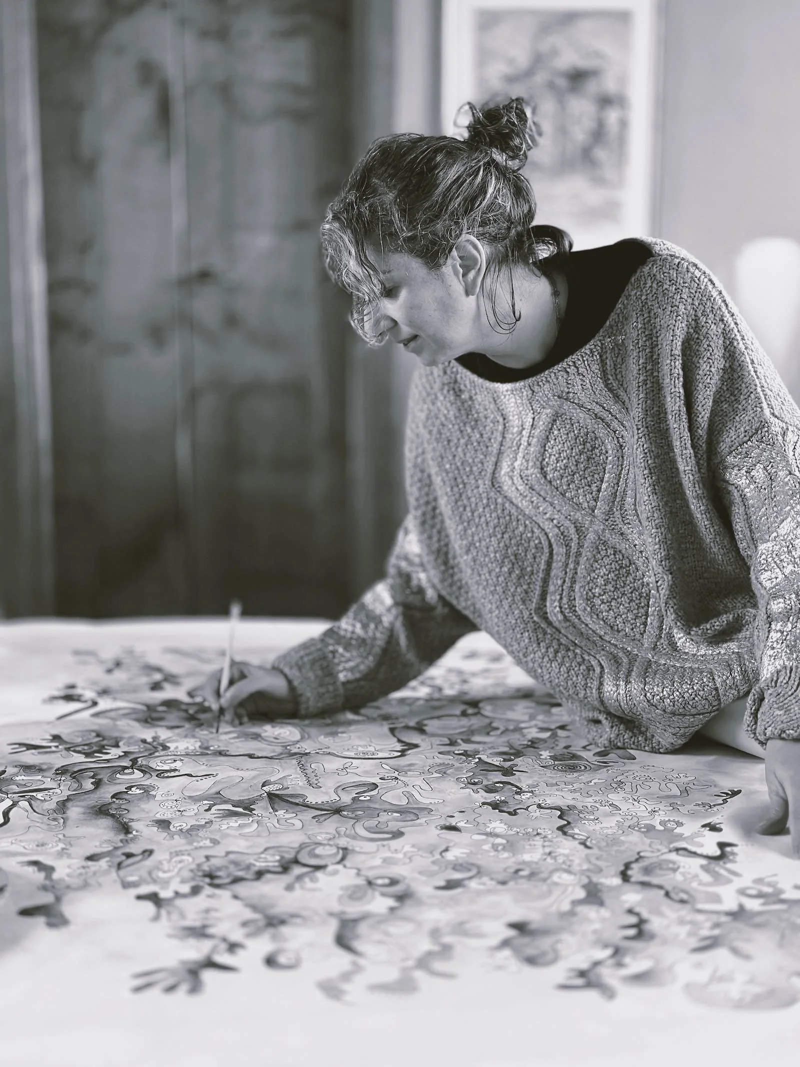 A woman with curly hair tied in a bun, wearing an oversized patterned sweater, is kneeling on a bed, painting an intricate design with a brush on a large canvas or paper spread out on the bed. The background features a wooden wall and framed artwork.