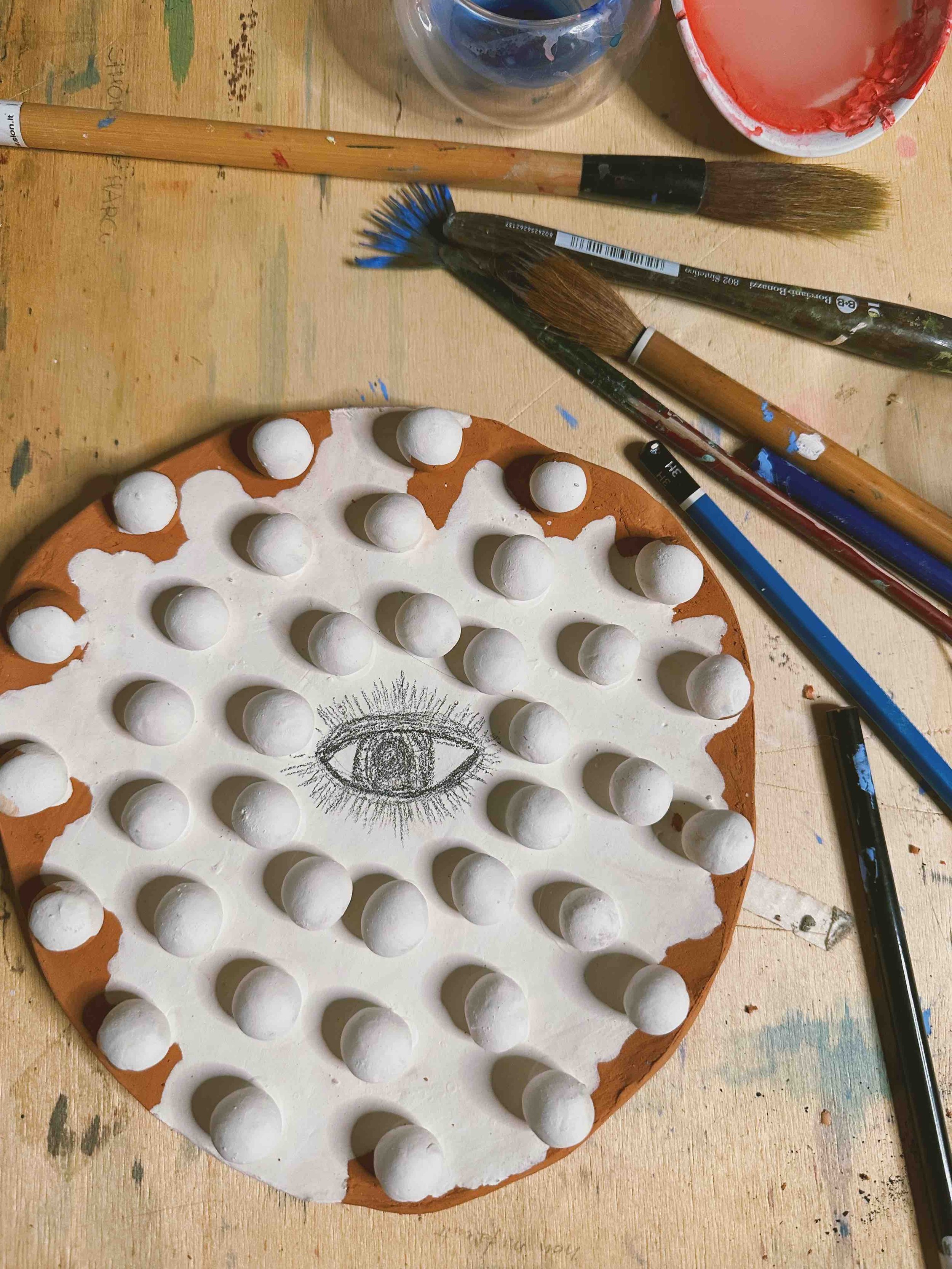 A wooden table with paintbrushes, paint cups, and a drawing of an eye surrounded by clay balls on a round clay surface.