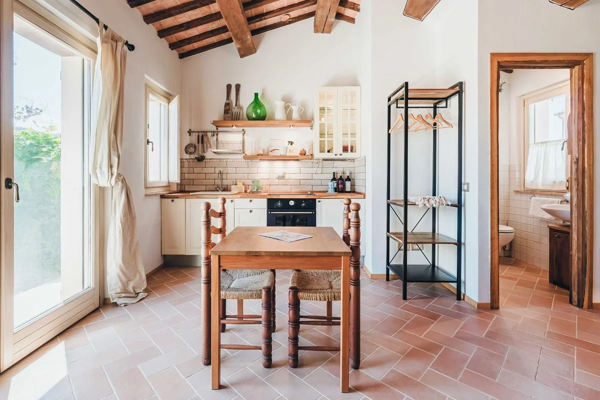 Bright kitchen with exposed wooden beam ceiling, white cabinetry, brick backsplash, small dining table with two chairs, open shelving with decorative items, black metal shelving unit, and a view into a small bathroom with a toilet and sink.