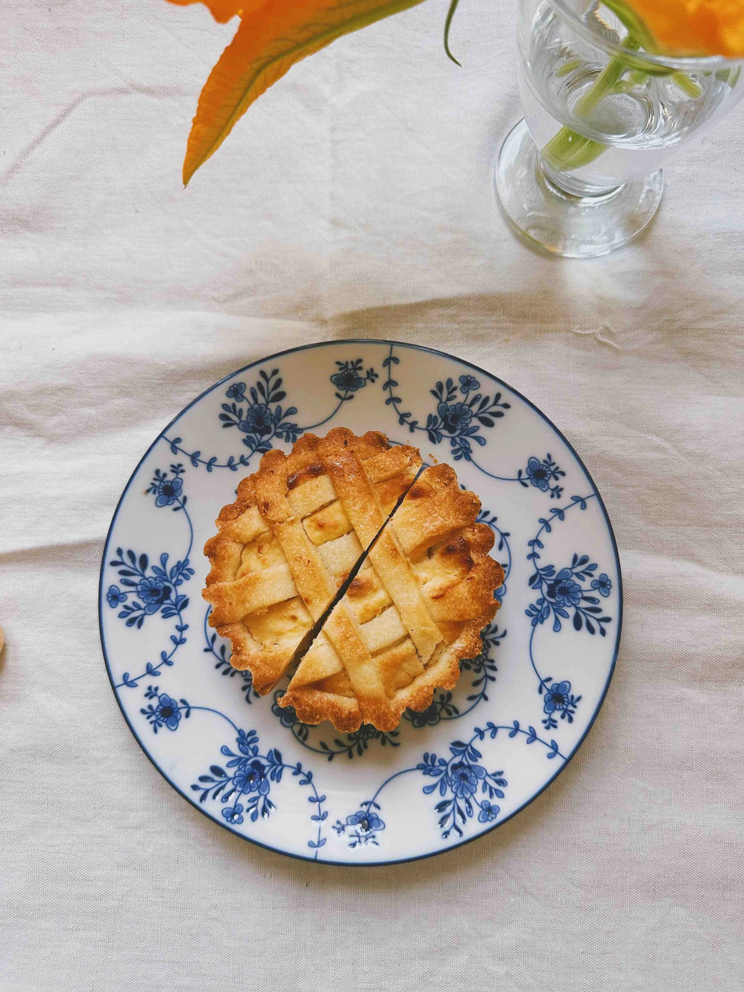 A slice of ricotta tart on a blue and white floral plate with a glass of water and an orange flower in the background.