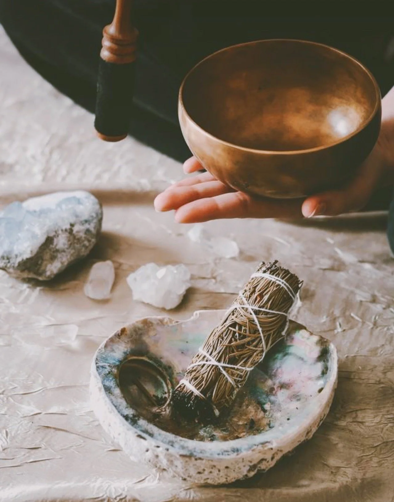 Francesca holding a singing bowl, with a collection of crystals and a bundle of sage on a stone platter, on a textured surface.