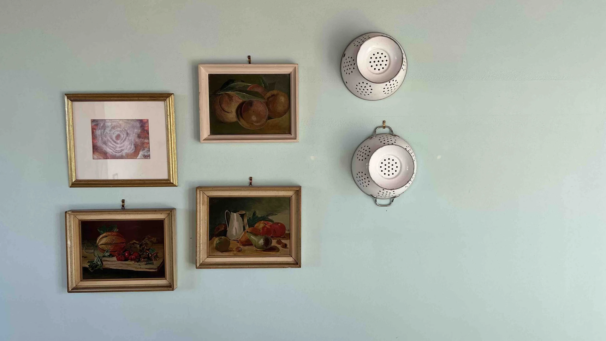 A mint green wall decorated with four framed still-life art pieces and two metal colanders. The frames are gold and wood, featuring 19th-20 century oil paintings of fruits and flowers.