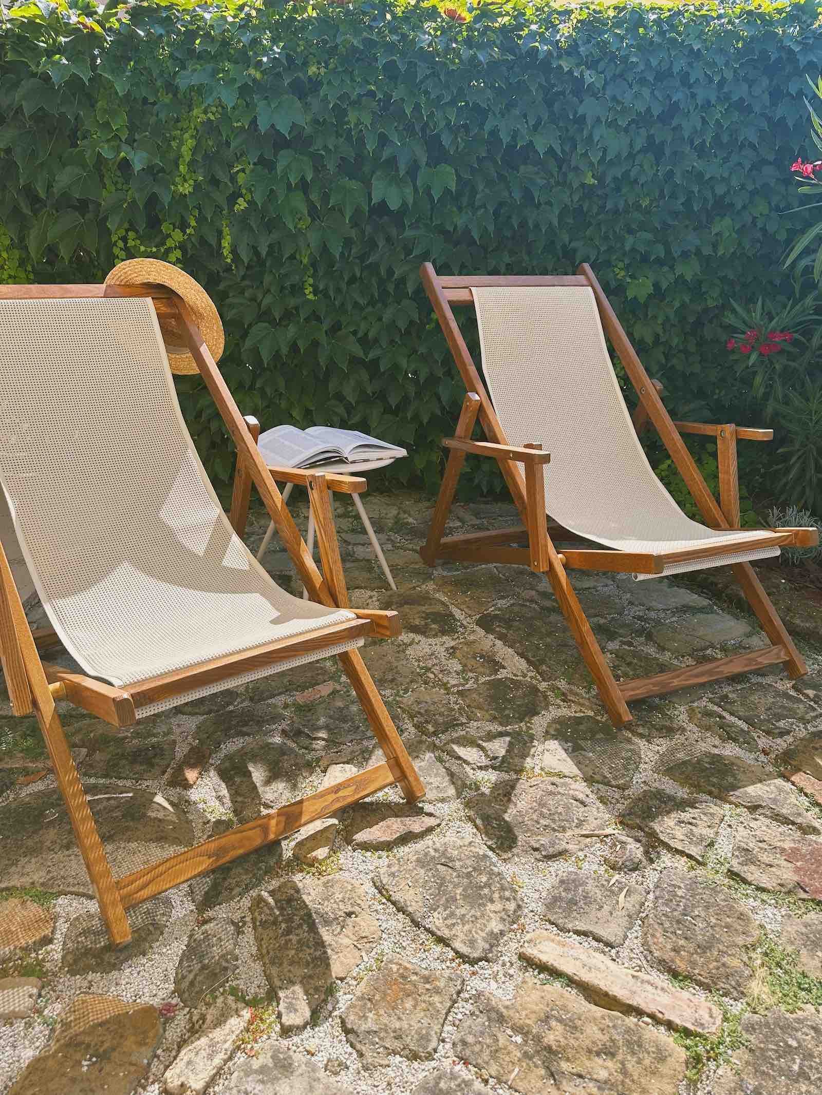 Two wooden outdoor lounge chairs with beige fabric, a straw sun hat hanging on one, and an open book on a small side table, set on a stone patio of Casa Janna private gardens with lush green foliage in the background.