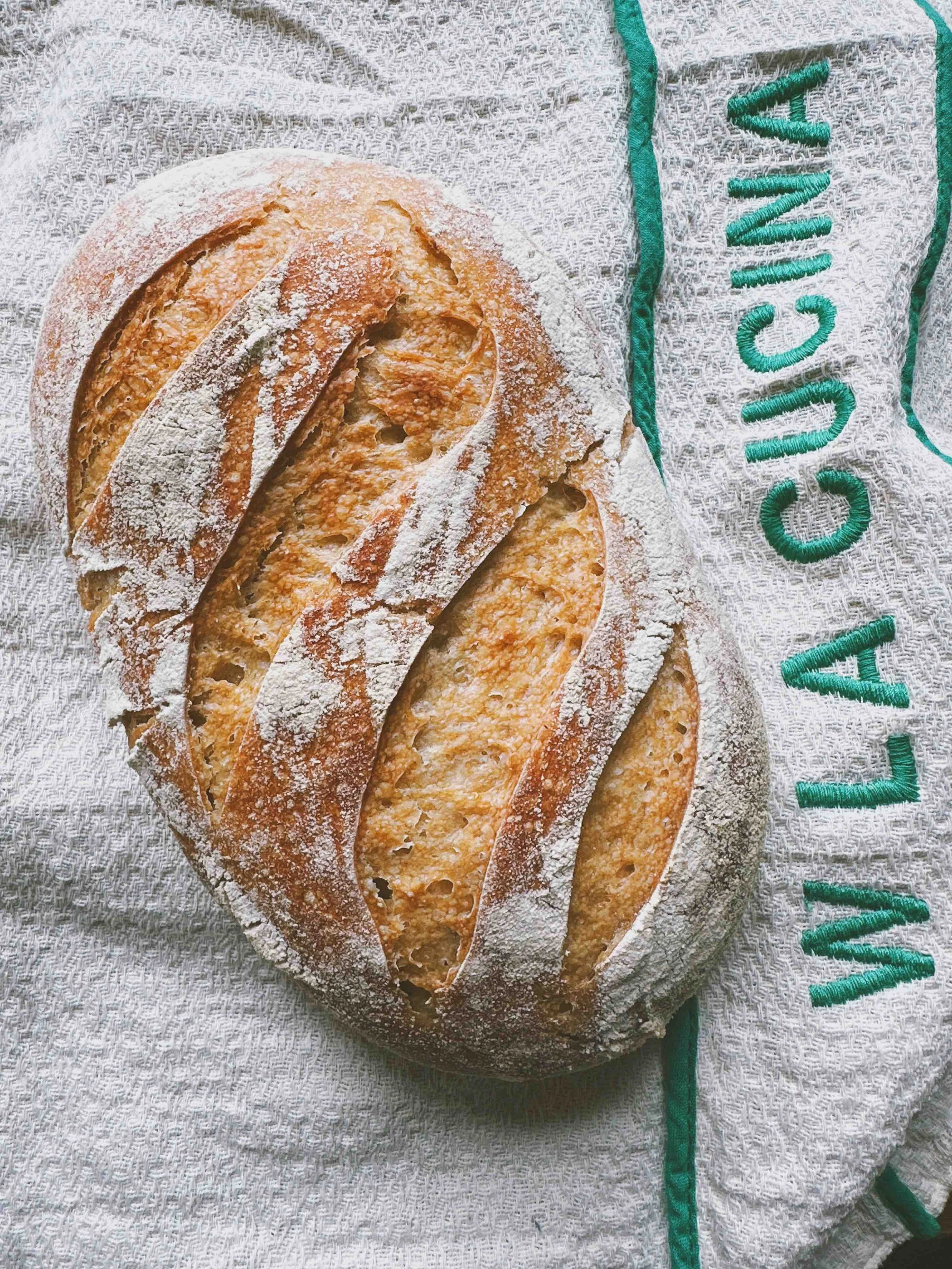 Freshly baked loaf of bread with a golden crust and dusted with flour, resting on a cloth with partially visible green embroidered text.