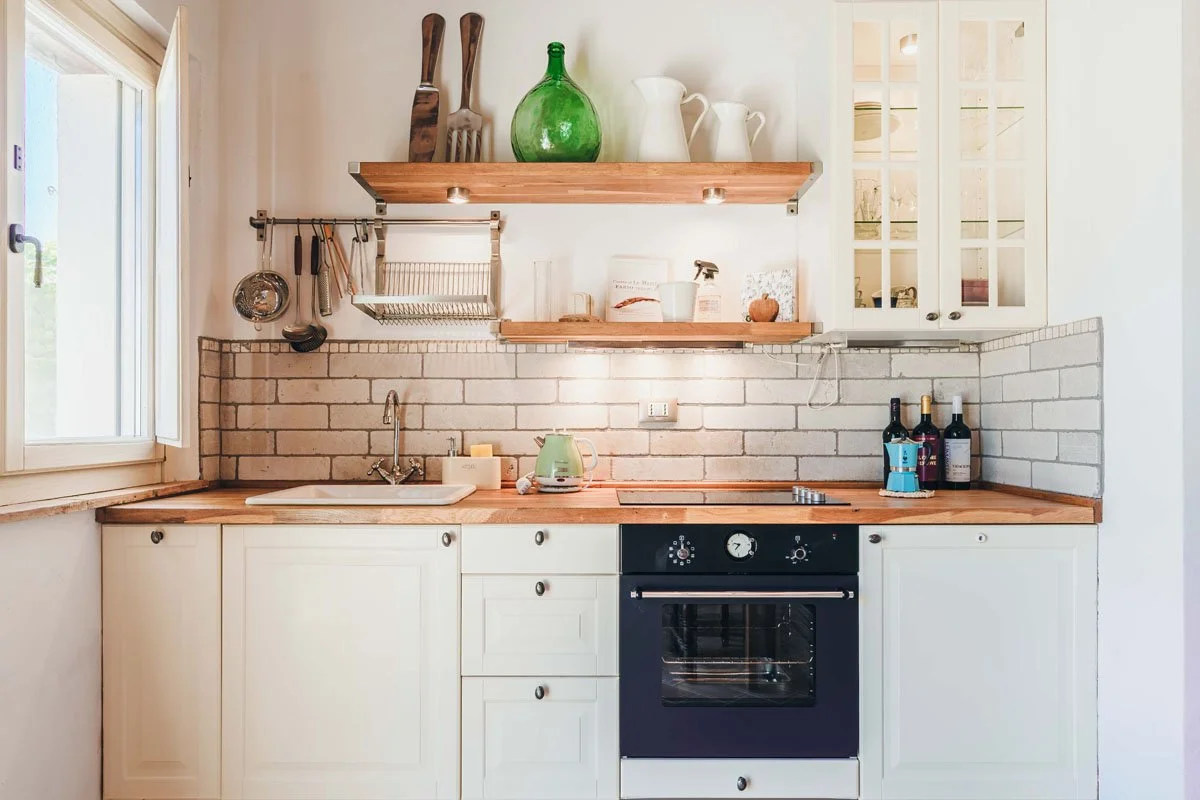 Kitchen with white cabinets, a wooden countertop, a window on the left, and a brick backsplash. There are open wooden shelves with dishes and bottles, a sink, a green kettle, and three wine bottles on the right.