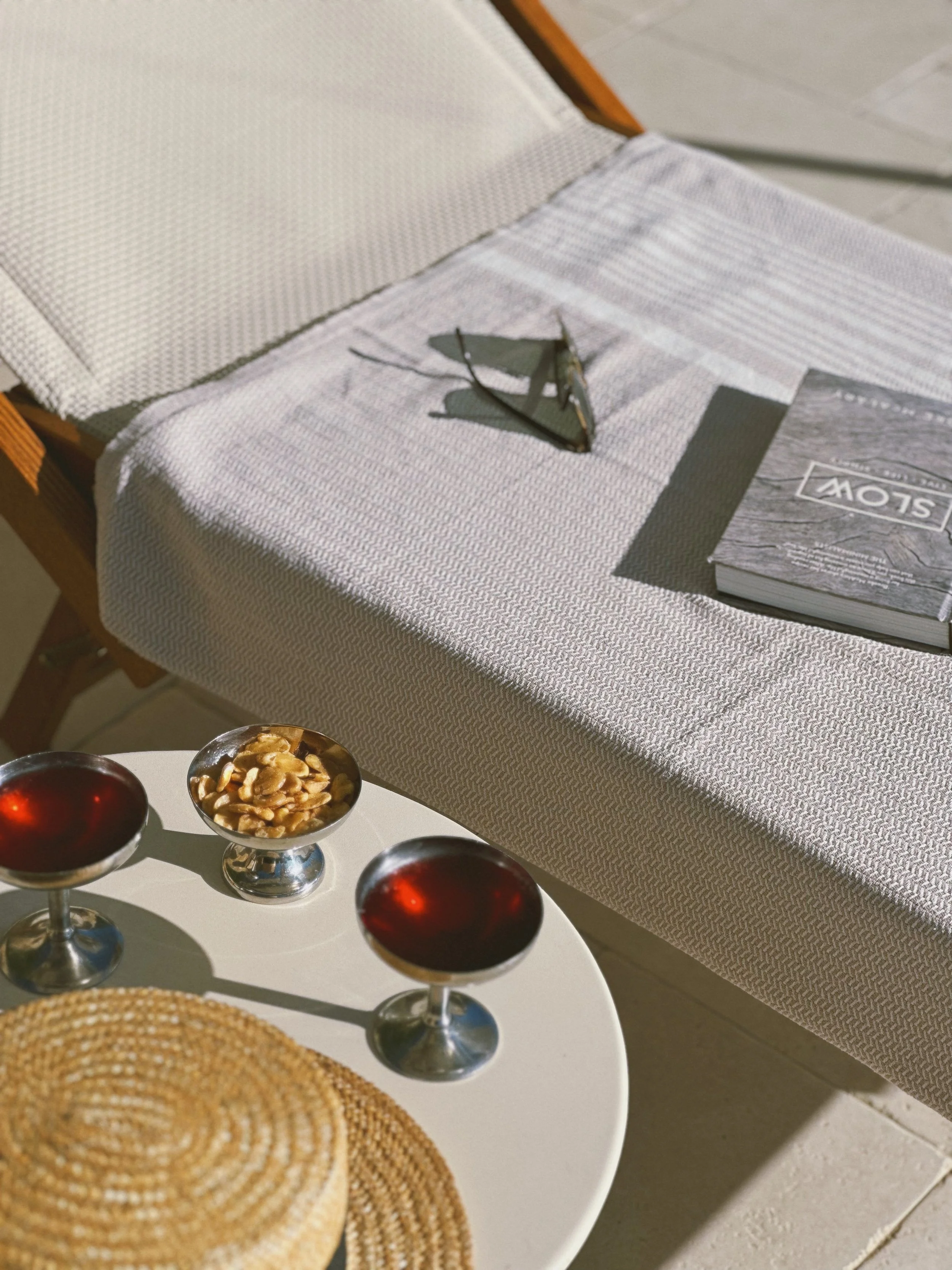 A white round table holding three glasses with dark red drinks, a small bowl of nuts, and a straw hat. In the background, a sunlit outdoor lounge chair has a folded towel, a magazine, and sunglasses resting on it.