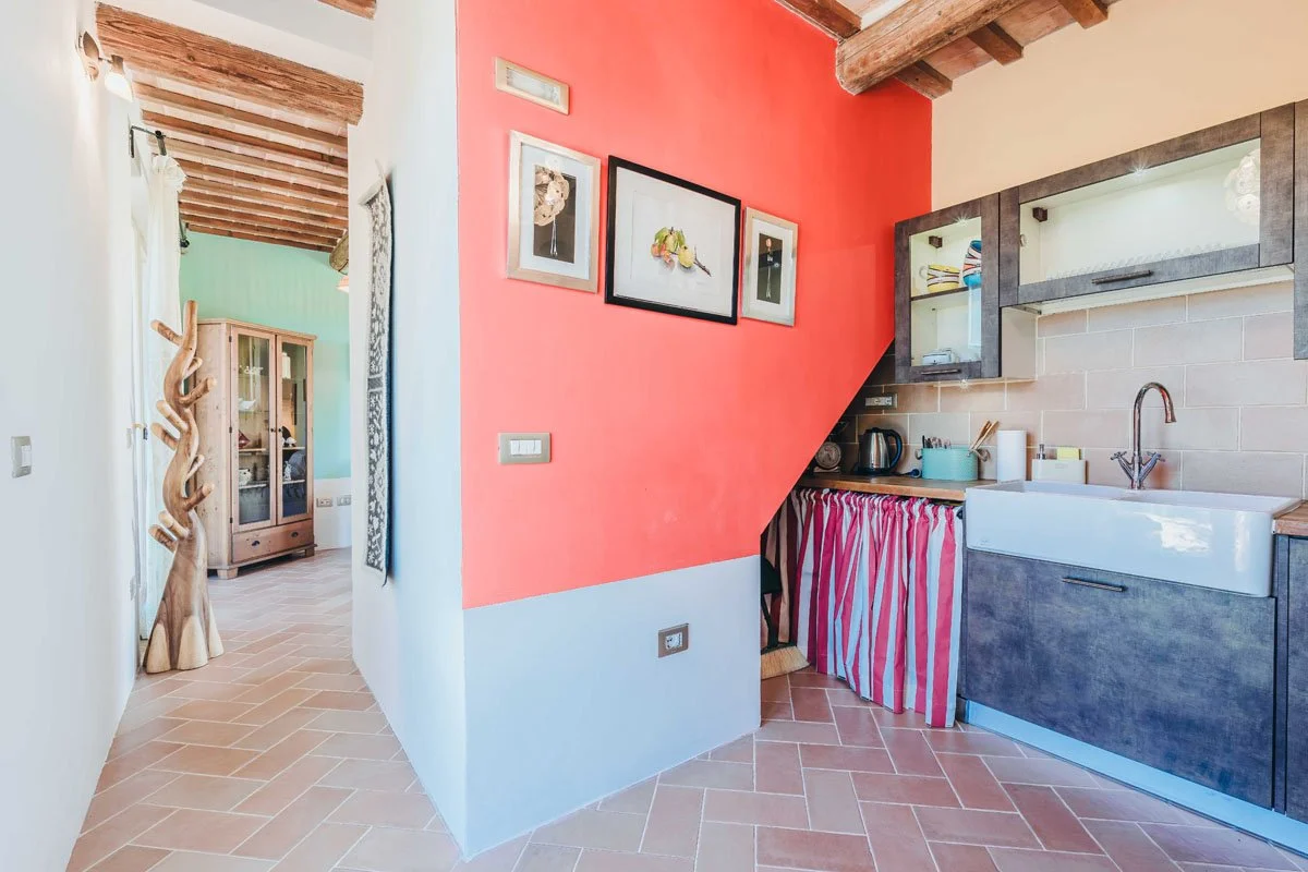 Kitchen interior at Casa Janna with mediterrean walls and a rustic style. There are picture frames on a wall, and a ceramic kitchen sink with dark wood cabinets and a striped curtain below. The floor is tiled with terracotta-colored tiles.