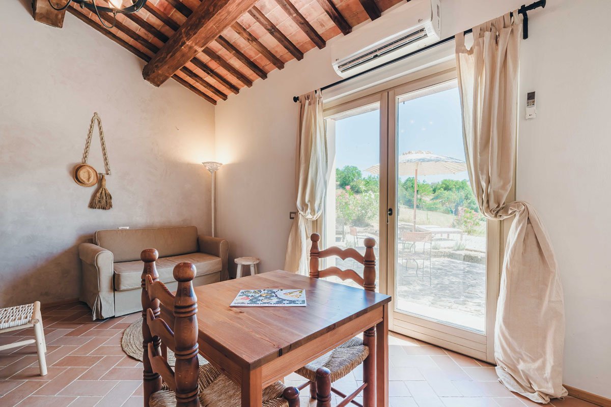 Cozy living room with a wooden dining table, wooden chairs, a beige sofa, a tall floor lamp, and large sliding glass door with cream curtains leading outside to patio with umbrella and outdoor furniture, wooden beams on the ceiling.
