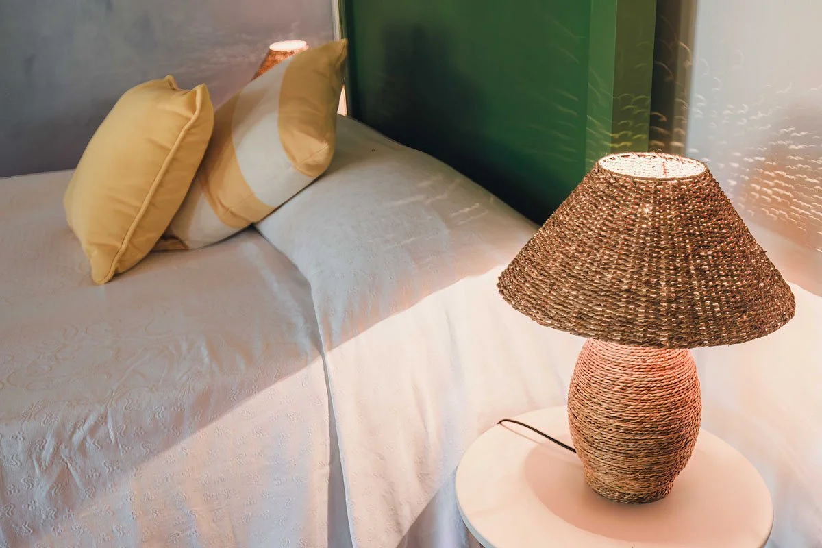 A bedside table with a woven wicker lamp and three pillows on a bed with a white textured bedspread, in a modern bedroom with green accents.