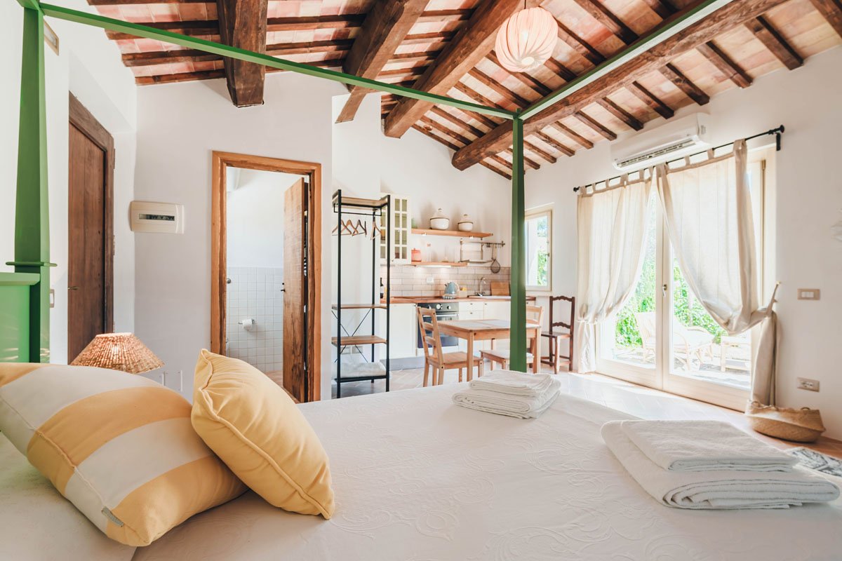 Bright bedroom with a bed, pillows, and blankets in the foreground, open kitchen with a dining table, and large windows with curtains letting in natural light. Wooden ceiling with exposed beams.