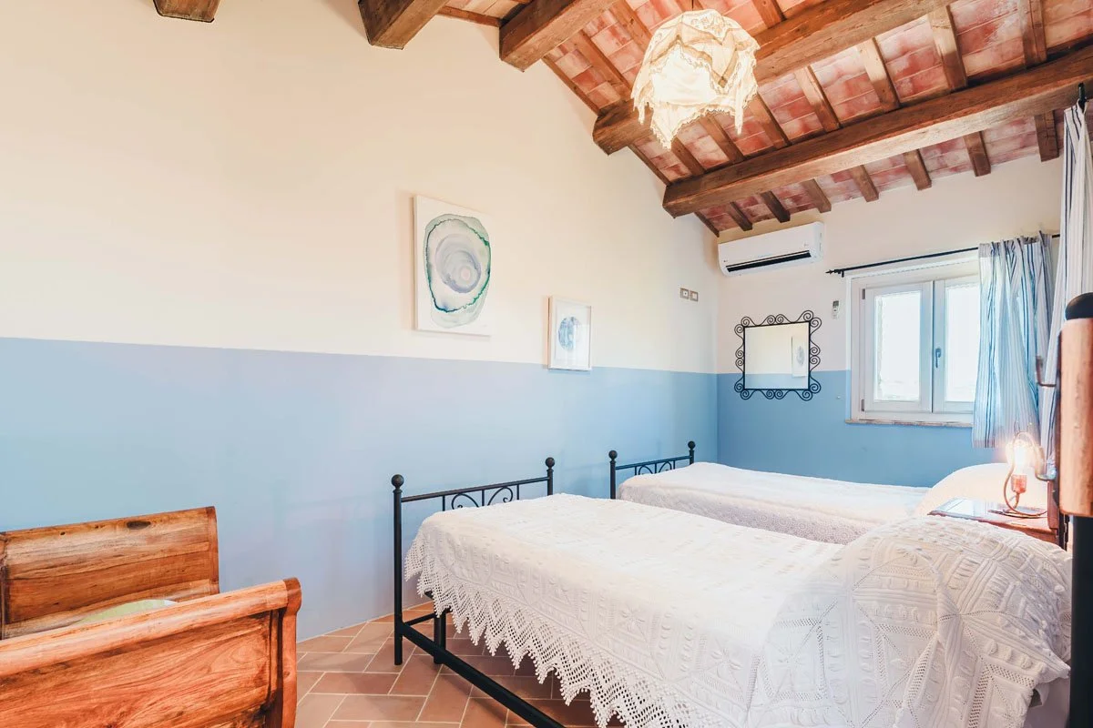 Bedroom at the Casa Janna Cottage with two iron beds, wooden ceiling beams, and natural light from a window