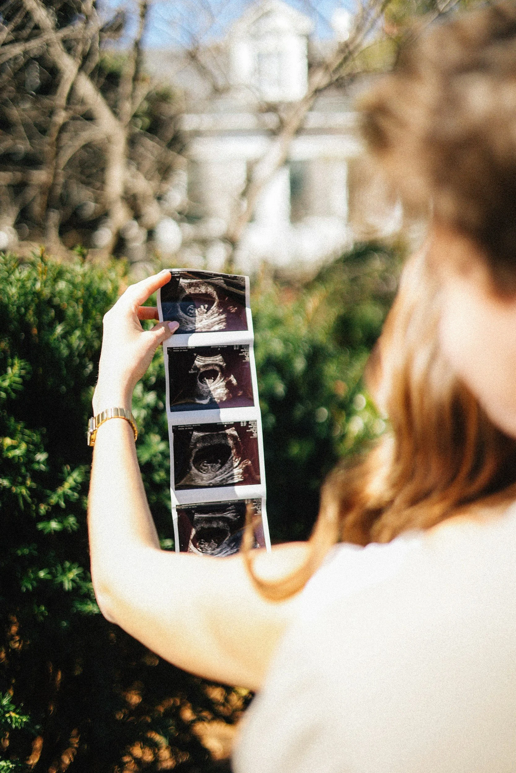 A woman holding and looking at ultrasound photos outdoors with greenery in the background.