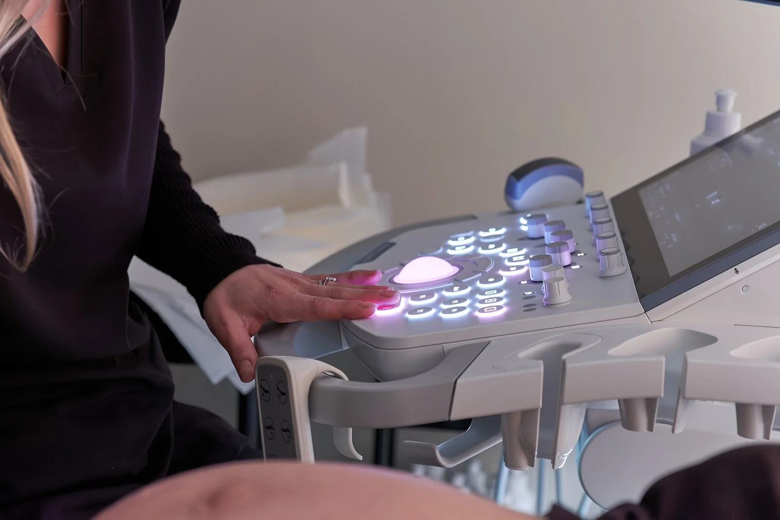 A person operating a medical ultrasound machine with a digital screen and control panel, in a clinical setting.