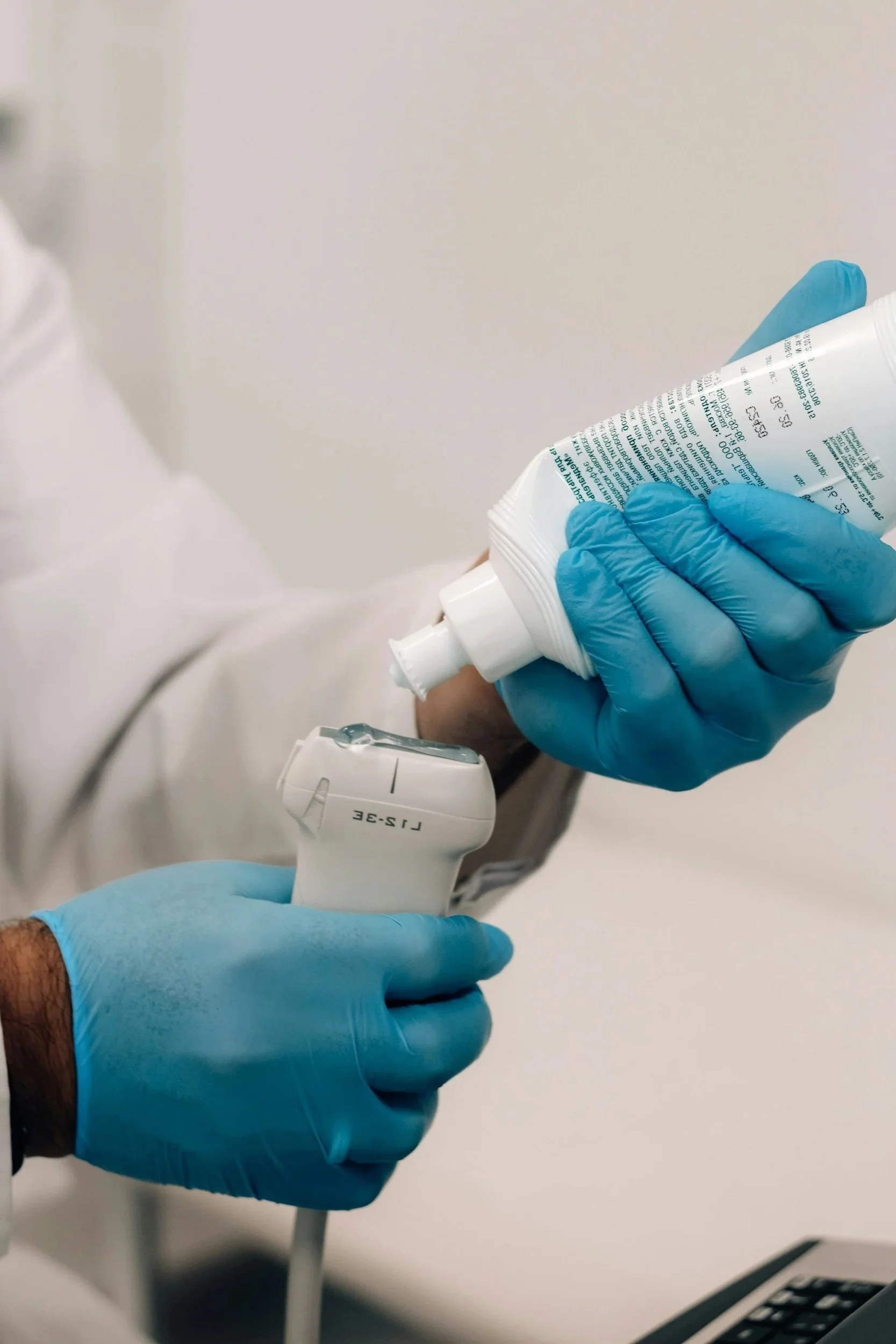 Person wearing blue gloves holding a tube of cream and a white device, with a partial view of a white coat and a white wall in the background.