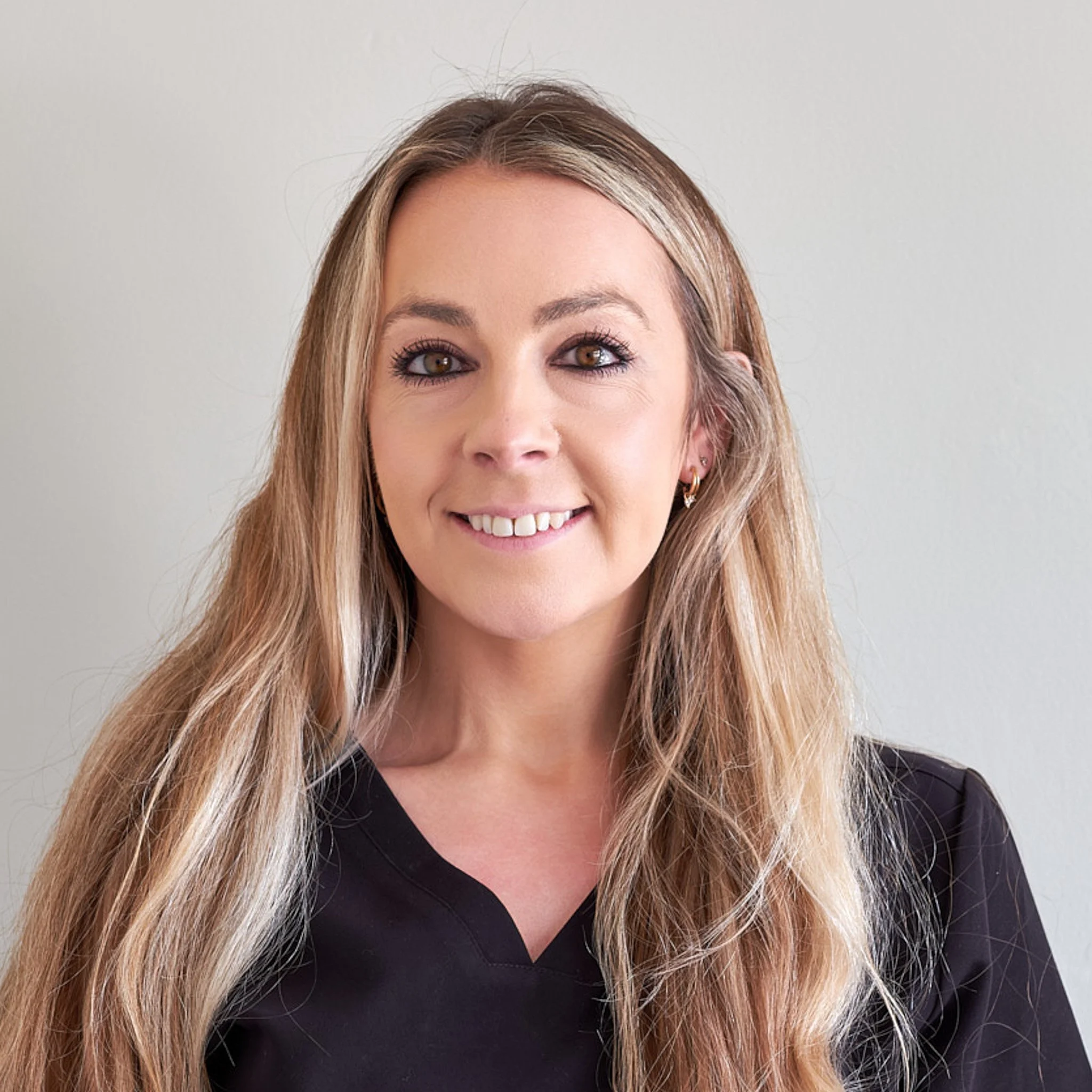 Close-up of a woman with long, wavy blonde hair, smiling, wearing a black V-neck top, with a plain light gray background.