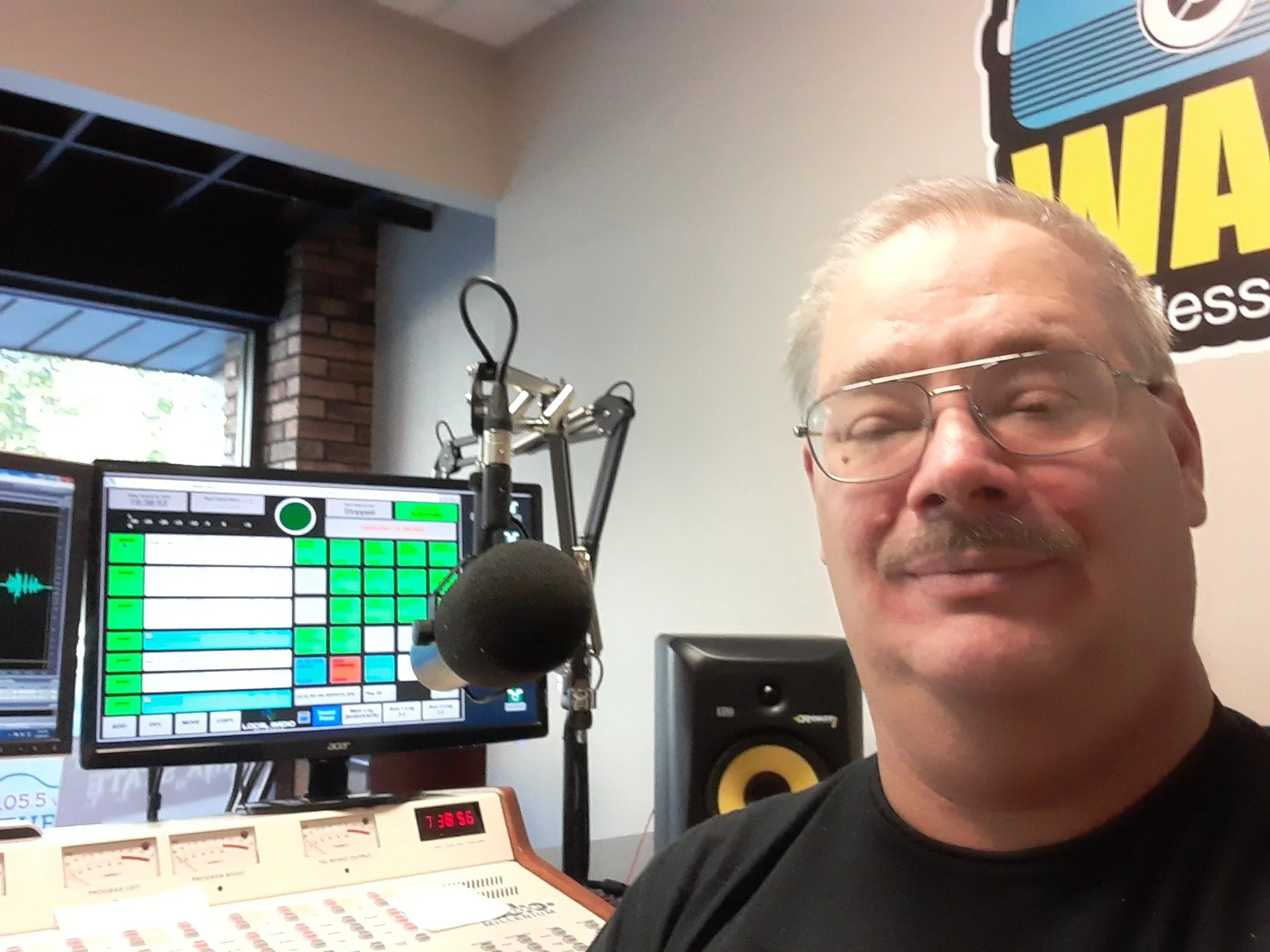 A man with glasses and a mustache smiling in a radio station studio, with a microphone, computer monitors, and audio equipment visible in the background.